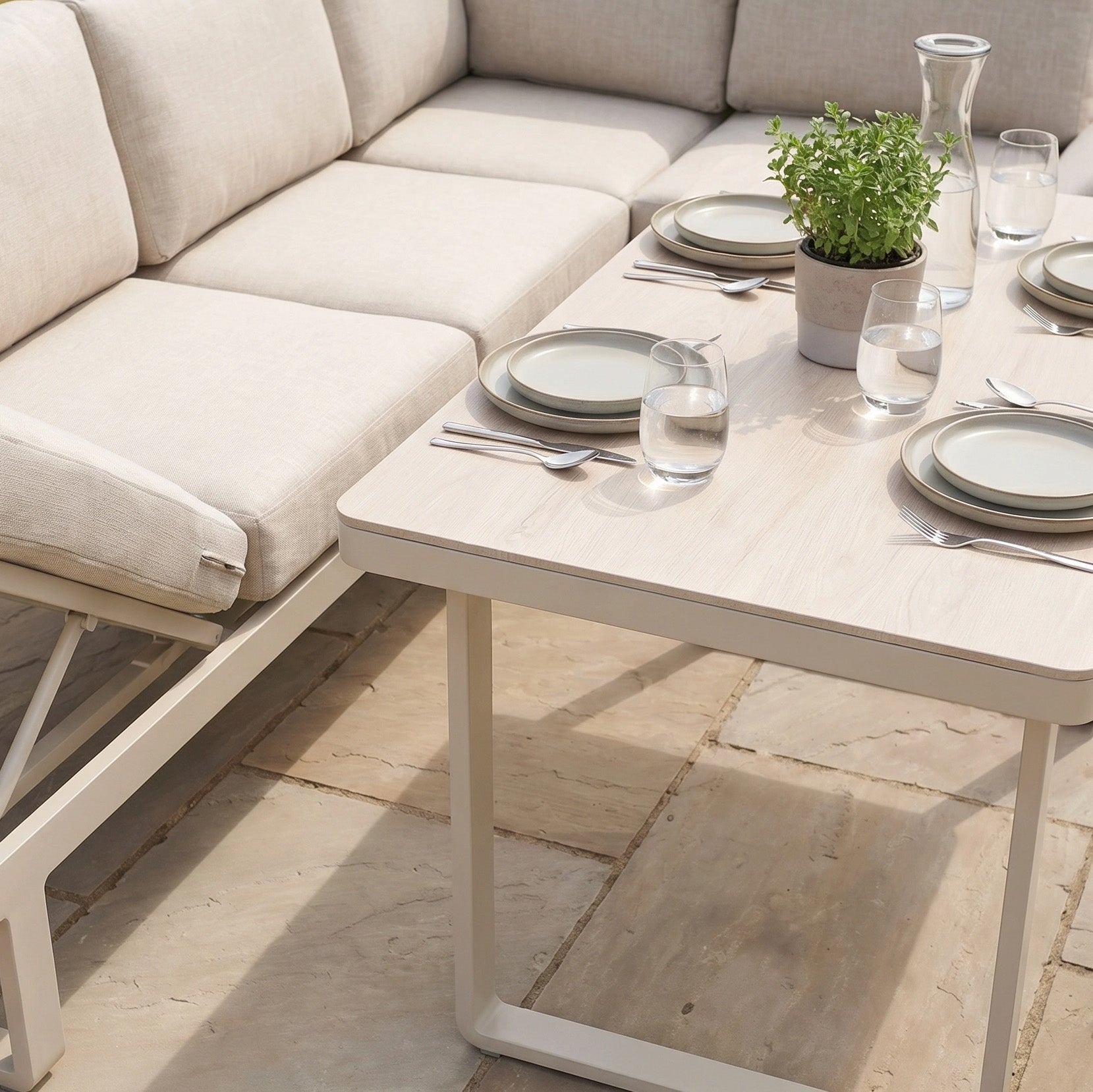 The Lyra Corner Dining Set in Latte features an outdoor patio table with plates, cutlery, glasses, a potted plant, a beige cushioned bench, and an elegant aluminium sofa.