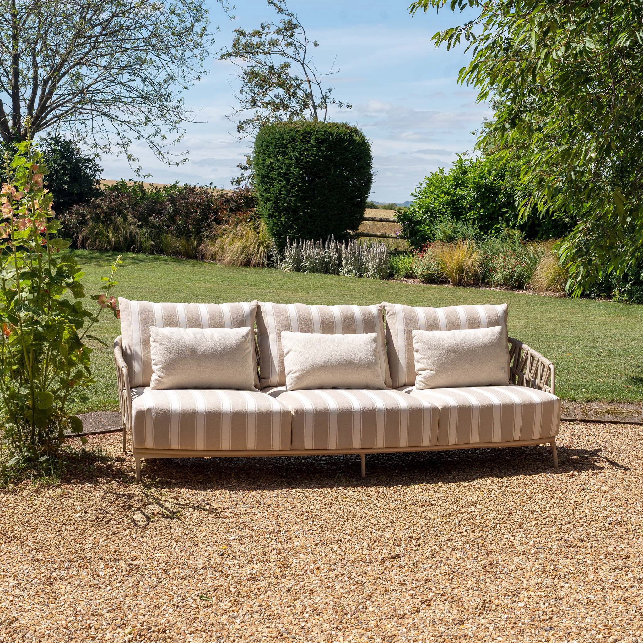 The Madeira 3 Seat Sofa in Striped Latte (Ex Display), ideal for garden furniture, sits on gravel in a garden with green grass and trees.