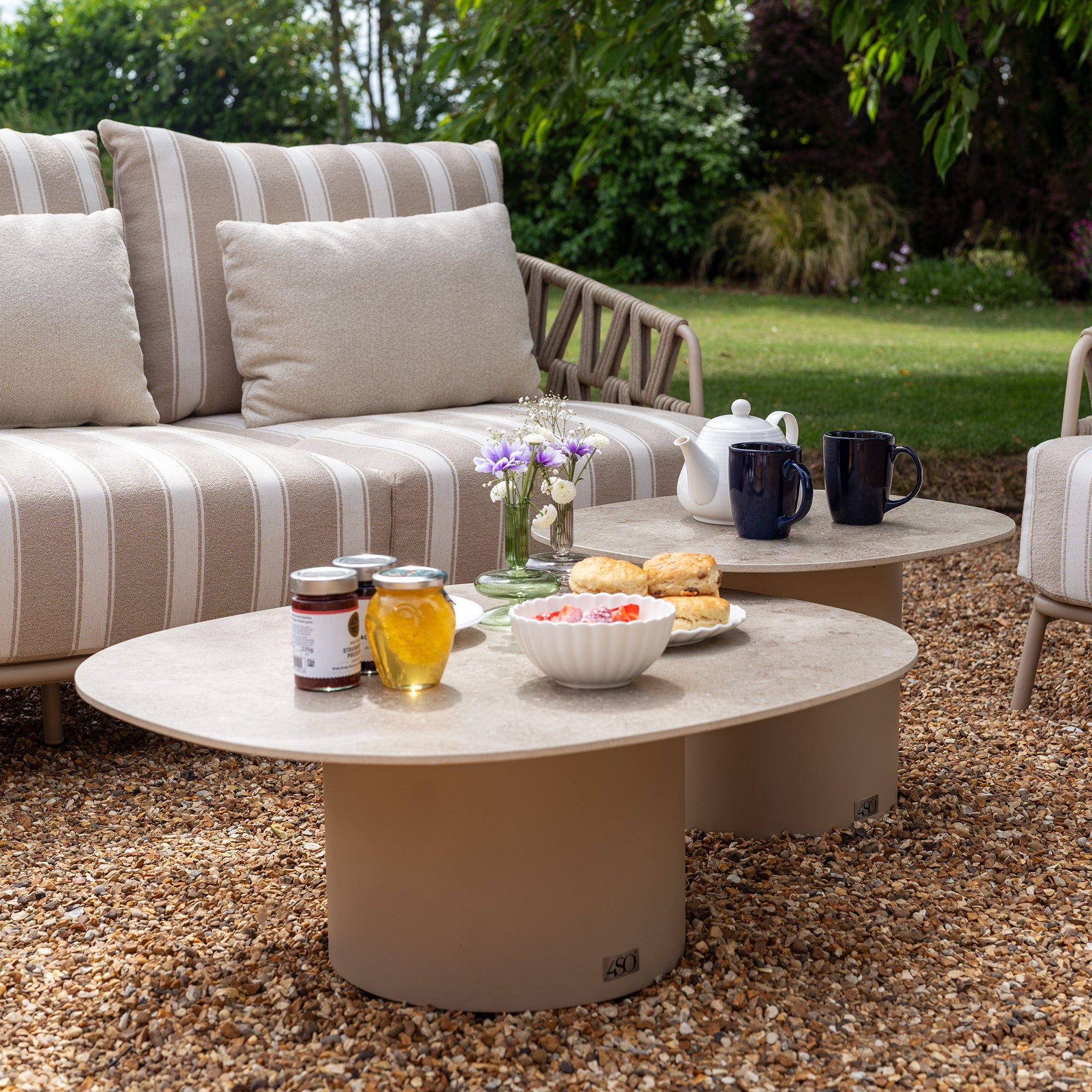 Two round tables with tea, scones, preserves, and flowers sit next to the Madeira 3 Seat Sofa Set in Striped Latte in a garden.