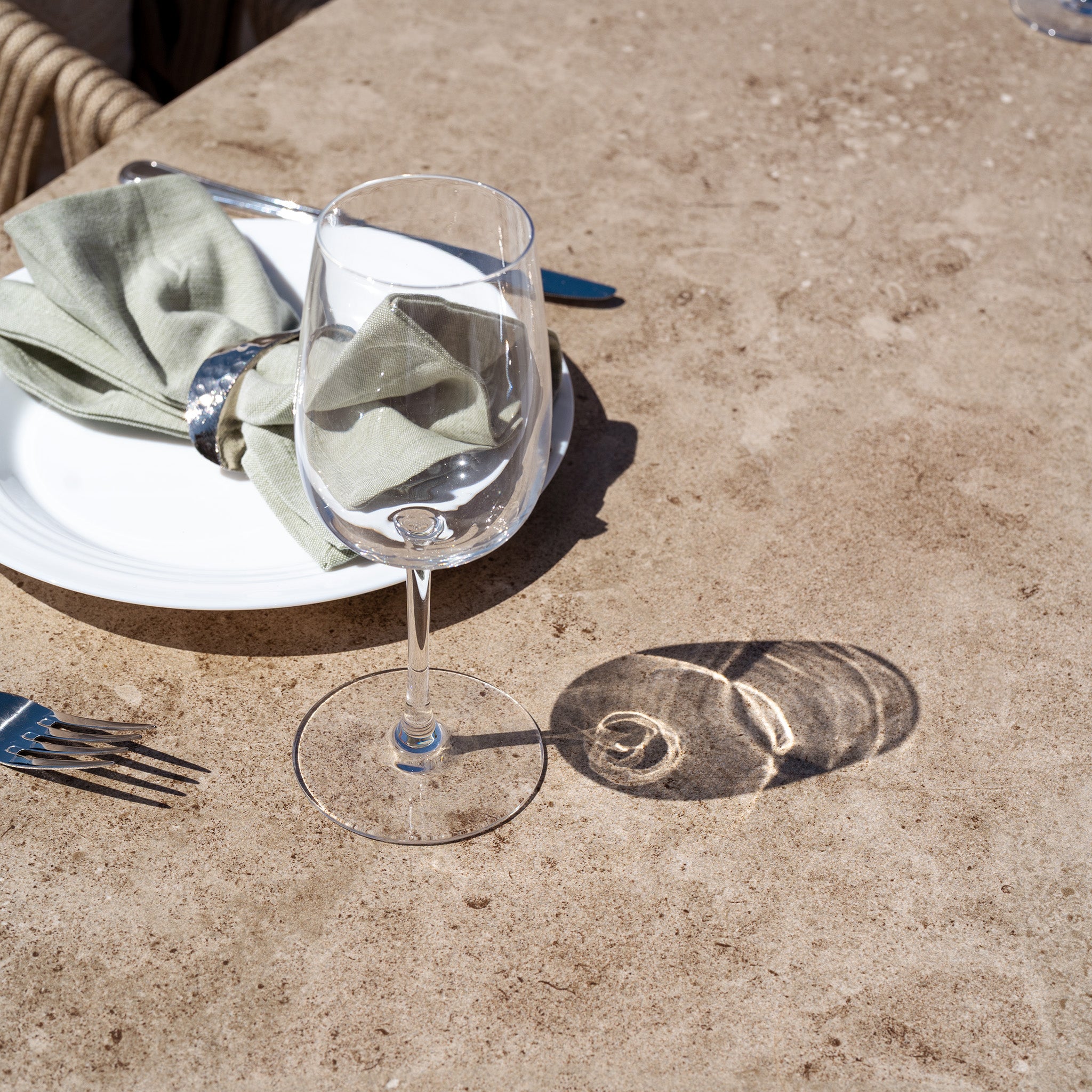 The Madeira 6 Seat Curved Rectangular Dining Set in Striped Latte is set on a stone table with an empty wine glass, cutlery, and a folded green napkin—perfect for an inviting alfresco meal.