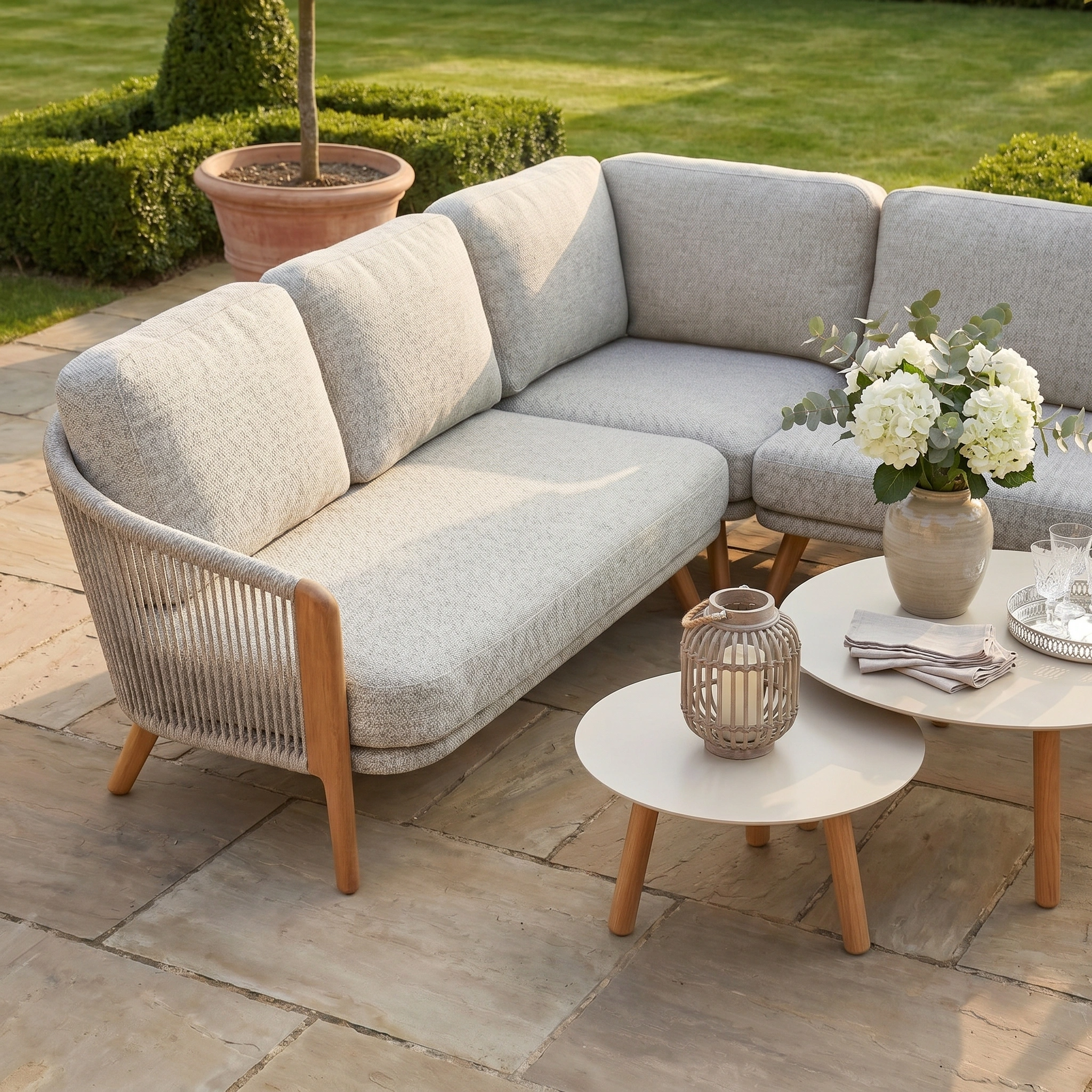 The Merano Corner Group Set with Coffee Table in Latte features an olefin rope weave, two round tables, flowers, and a lantern on a stone patio in a garden.