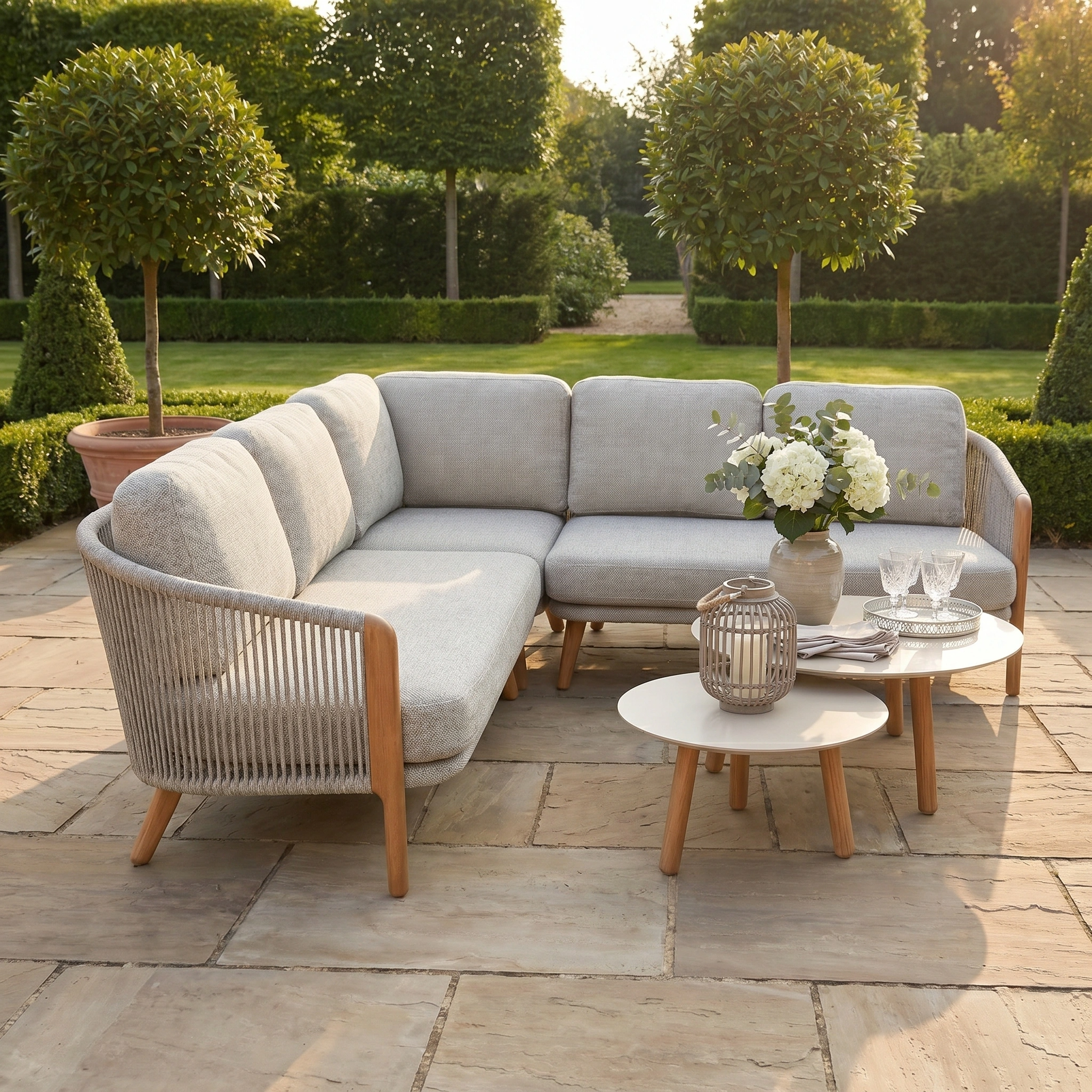 Outdoor patio with a Merano Corner Group Set with Coffee Table in Latte, featuring olefin rope weave, round tables, and potted trees on stone tiles in a garden setting.