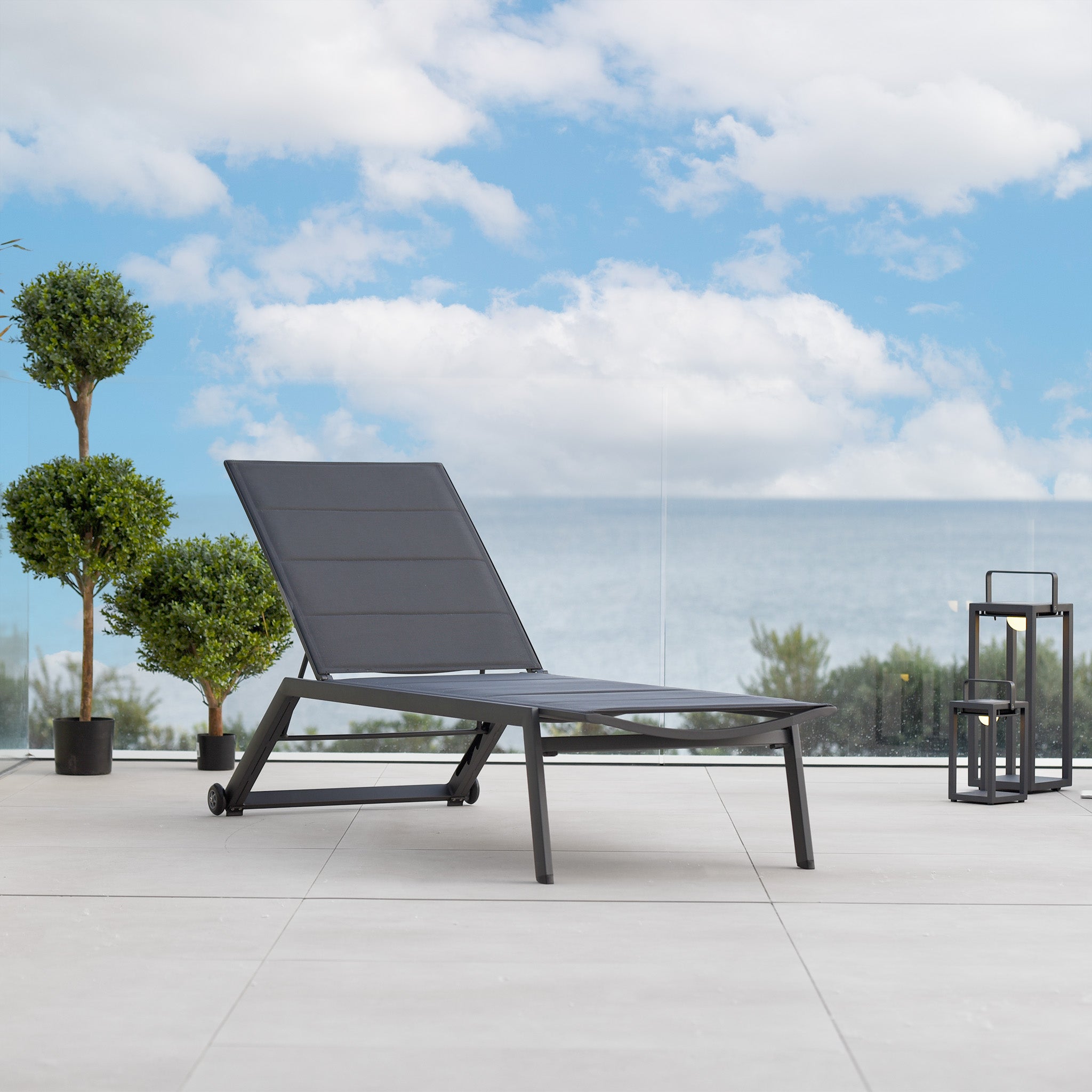 A modern charcoal Monaco Padded Sunlounger on a patio overlooking the ocean, with potted plants and lanterns nearby under a blue sky.