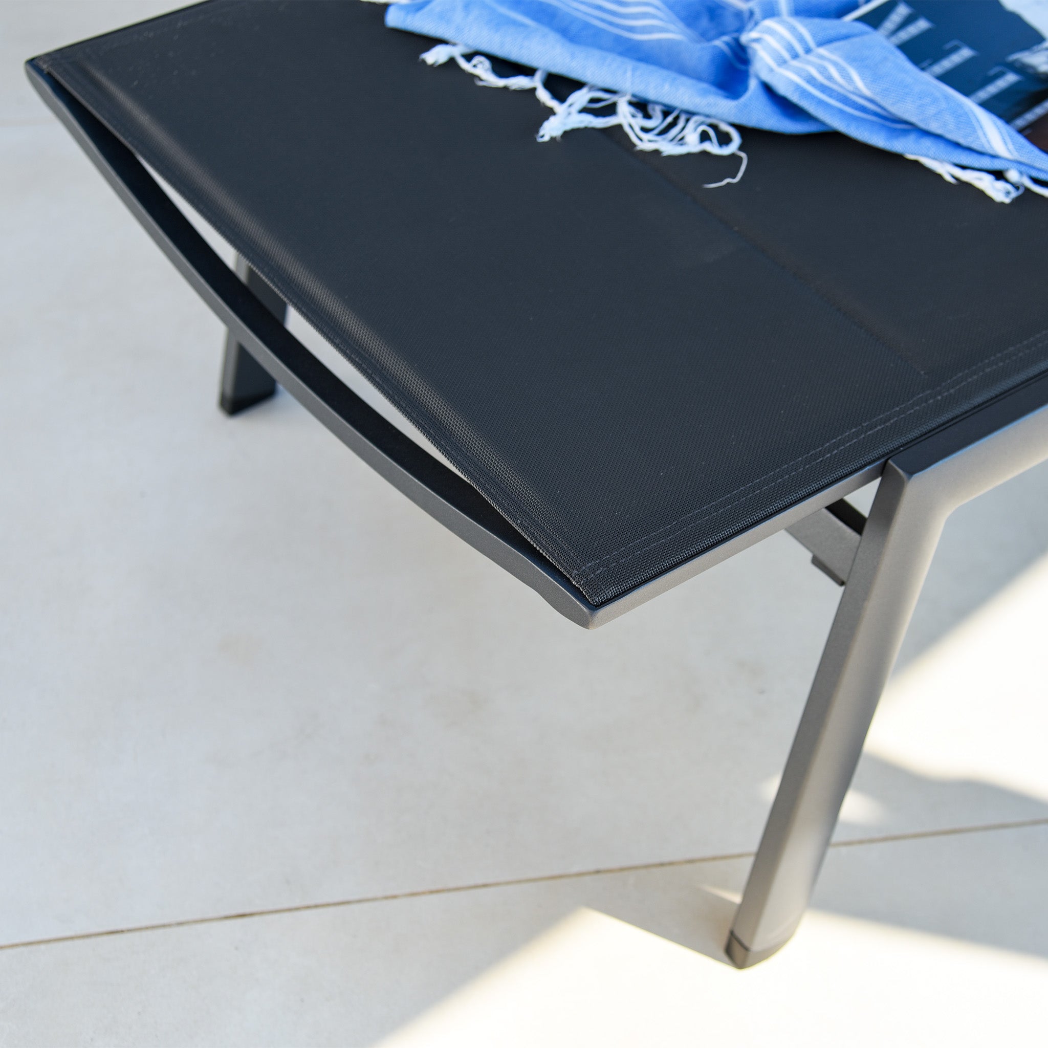 A Monaco Padded Sunlounger in Charcoal, featuring tear-resistant fabric, rests on a tiled patio with a blue and white striped towel draped over its black aluminium backrest.
