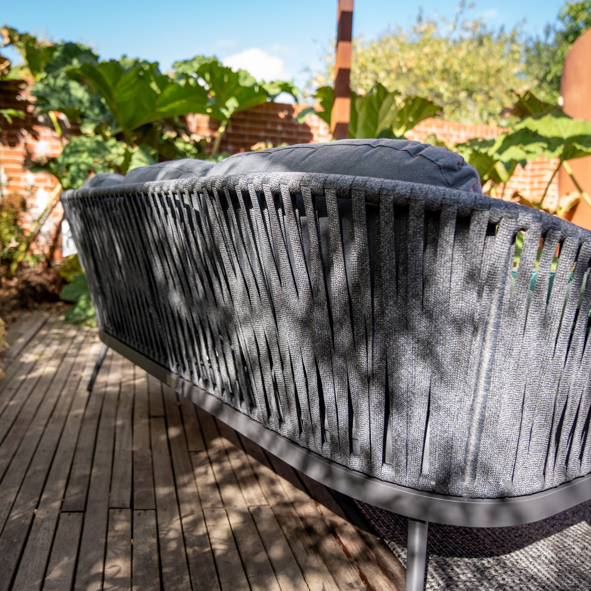 The Harbour Lifestyle Monterrey 3 Seat Rope Sofa Set with Rising Firepit Table in Grey features a woven backrest, elegantly positioned on a wooden deck amidst lush greenery and dappled sunlight. Spend your evenings by the built-in firepit, relaxing in sophisticated style.