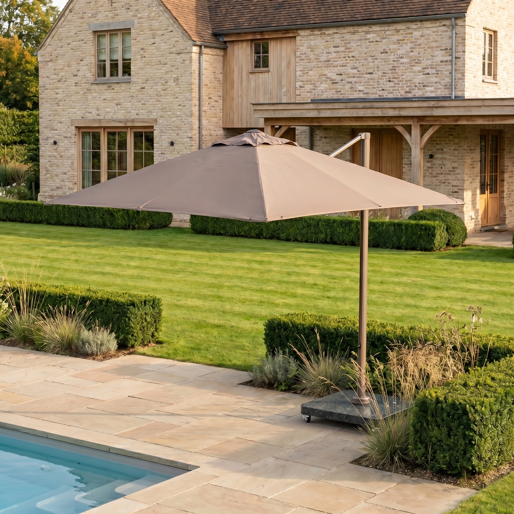 The Nexus T2 Premium 3m Square Cantilever Parasol in Deep Taupe with 90kg Granite Base is set beside stylish garden furniture on a stone patio overlooking a landscaped garden and swimming pool.