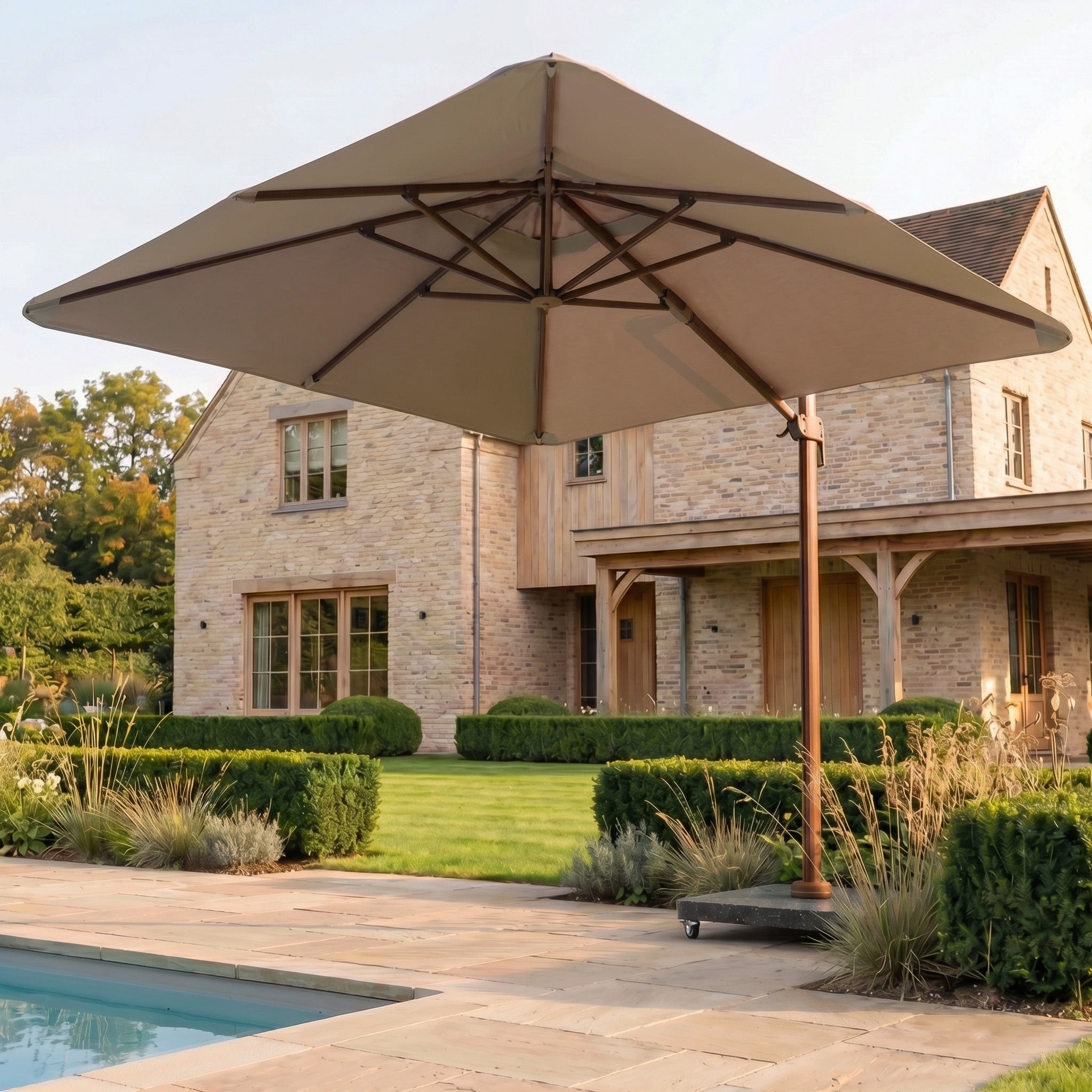 The Nexus T2 Premium 3m Square Cantilever Parasol in Deep Taupe with a 90kg Granite Base shades elegant garden furniture by the pool in front of a stone house with lush lawn and bushes.