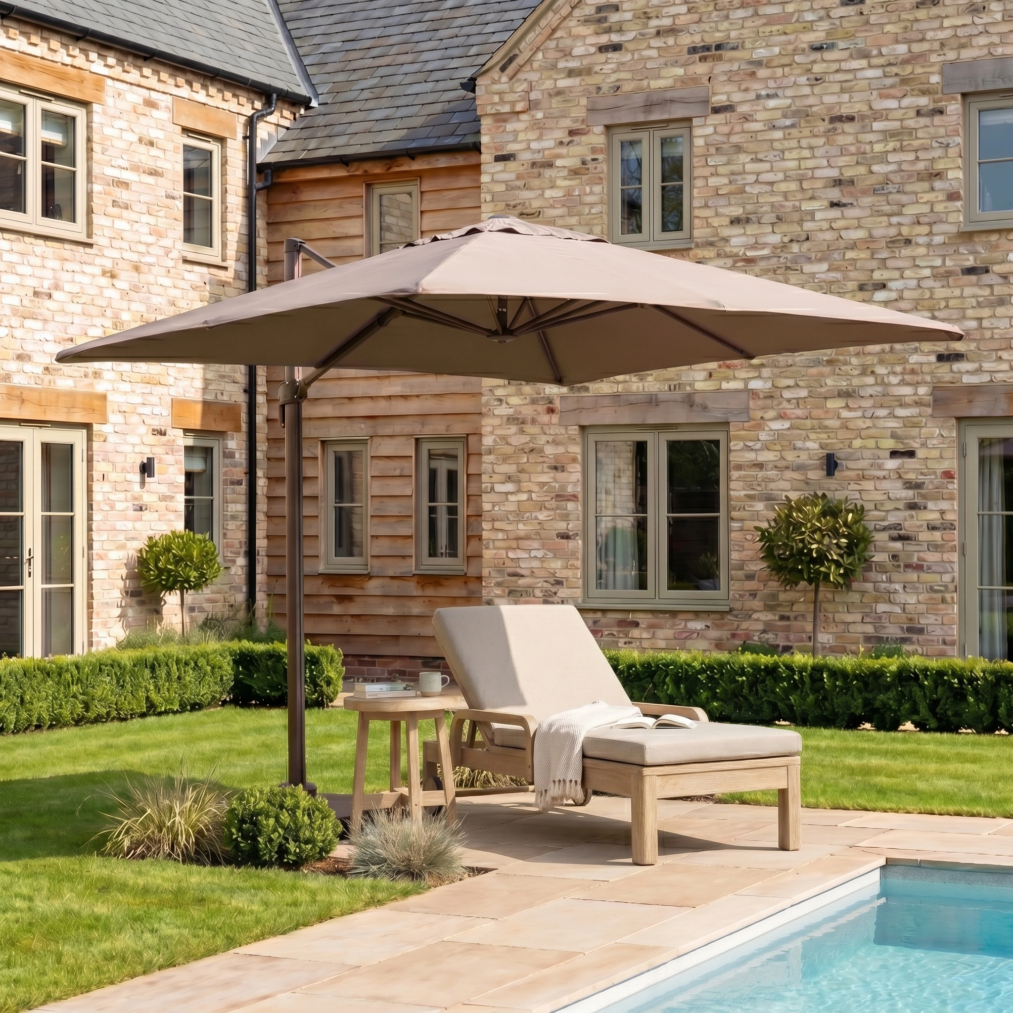 A wooden lounge chair and garden furniture with the Nexus T2 Premium 3m Square Cantilever Parasol in Deep Taupe with 90kg Granite Base by a pool in a sunny backyard next to a brick and wood house.