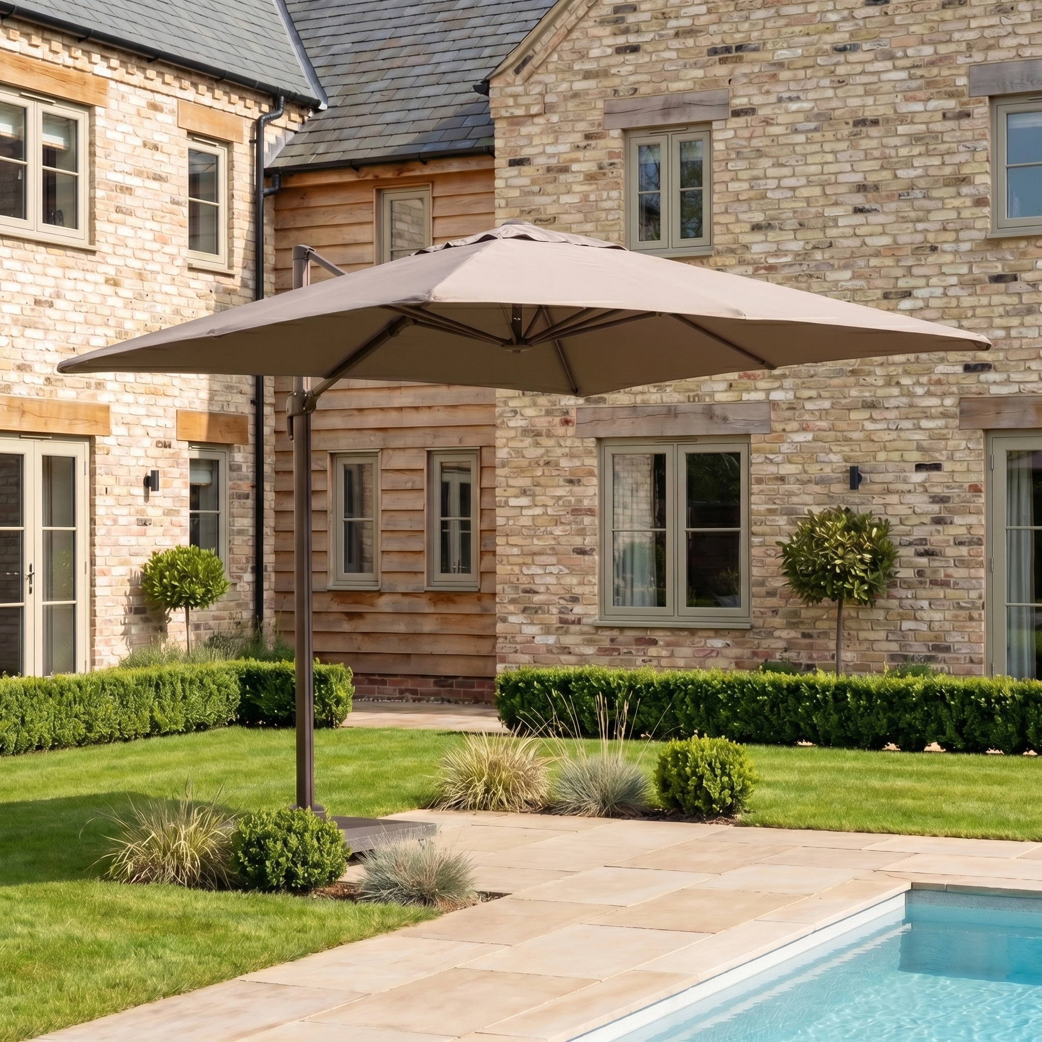 The Nexus T2 Premium 3m Square Cantilever Parasol in Deep Taupe with a 90kg granite base shades the garden of a brick house, perfectly complementing the stylish poolside furniture.