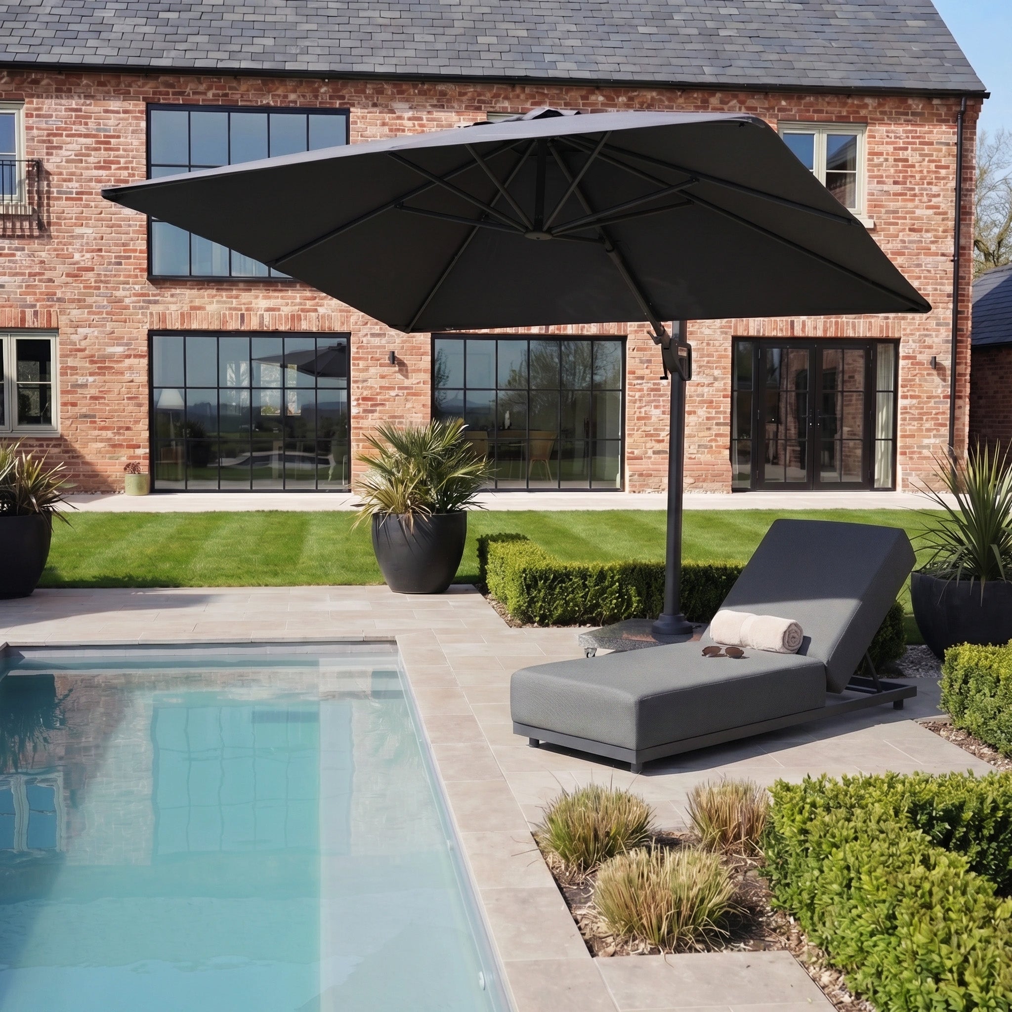 The gray lounge chair and Nexus T2 Premium 3m Square Cantilever Parasol in Black with 90kg Granite Base stand by a pool, set against a brick house and manicured garden in the background.