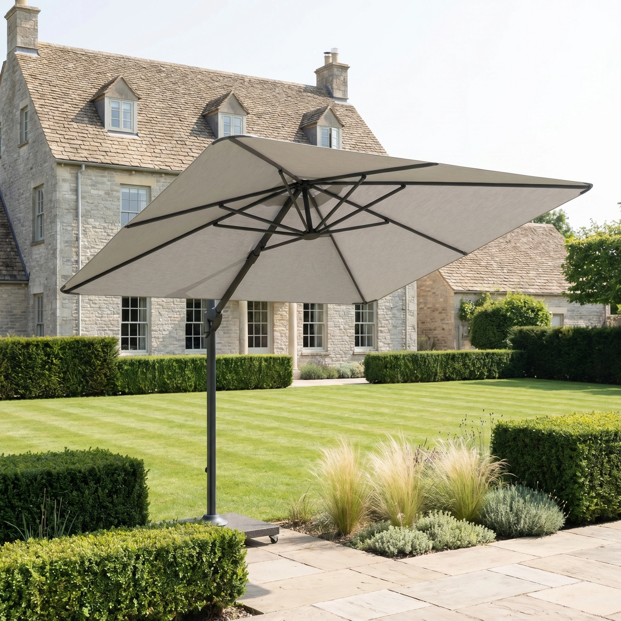 The Nexus T2 Premium 3m Square Cantilever Parasol in Manhattan Grey, with a 90kg granite base, features 360-degree rotation and stands in a manicured garden before a stone house.