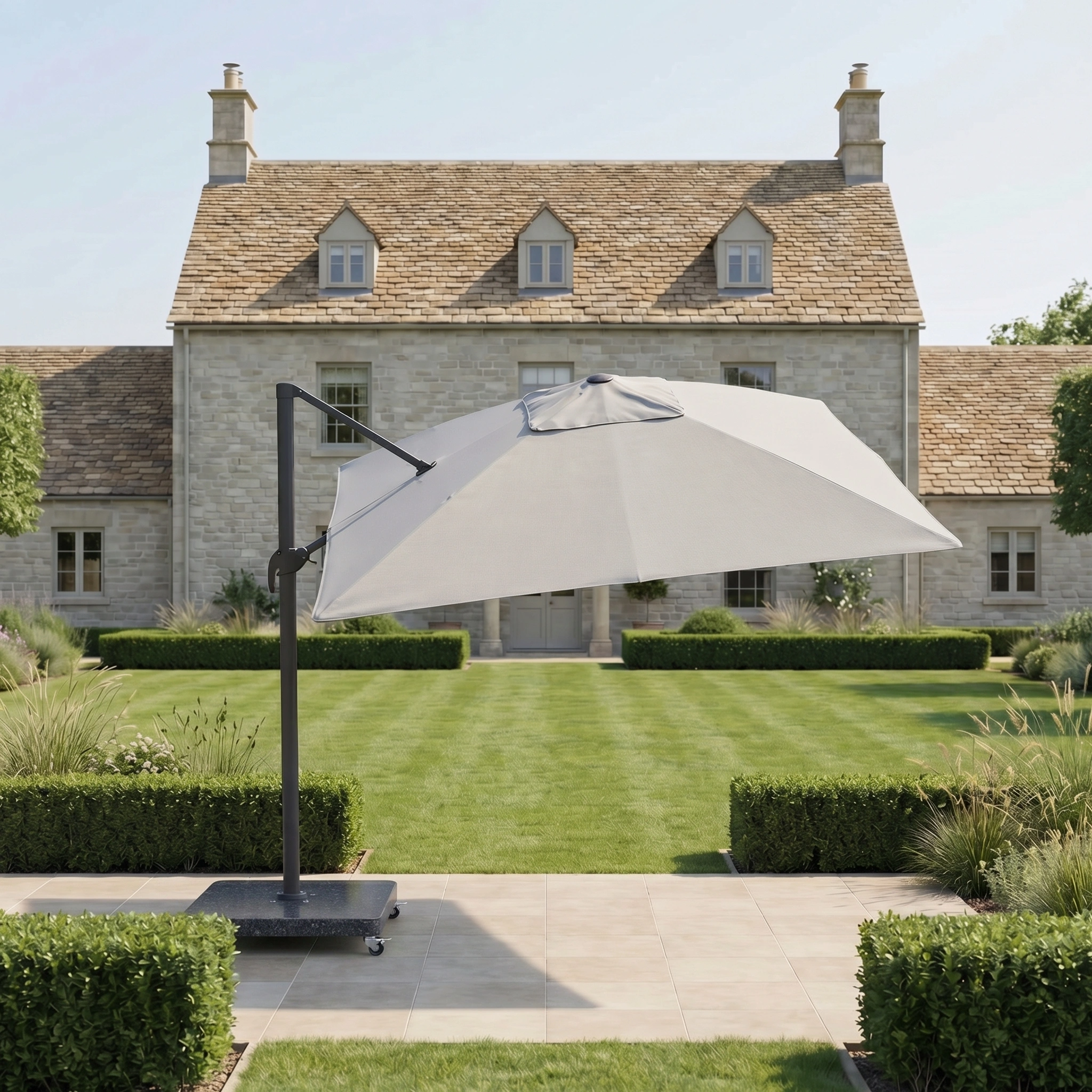The Nexus T2 Premium 3m Square Cantilever Parasol in Manhattan Grey, paired with a 90kg granite base, stands open in a manicured garden, shading the area before a charming stone house with its smooth 360-degree rotation.