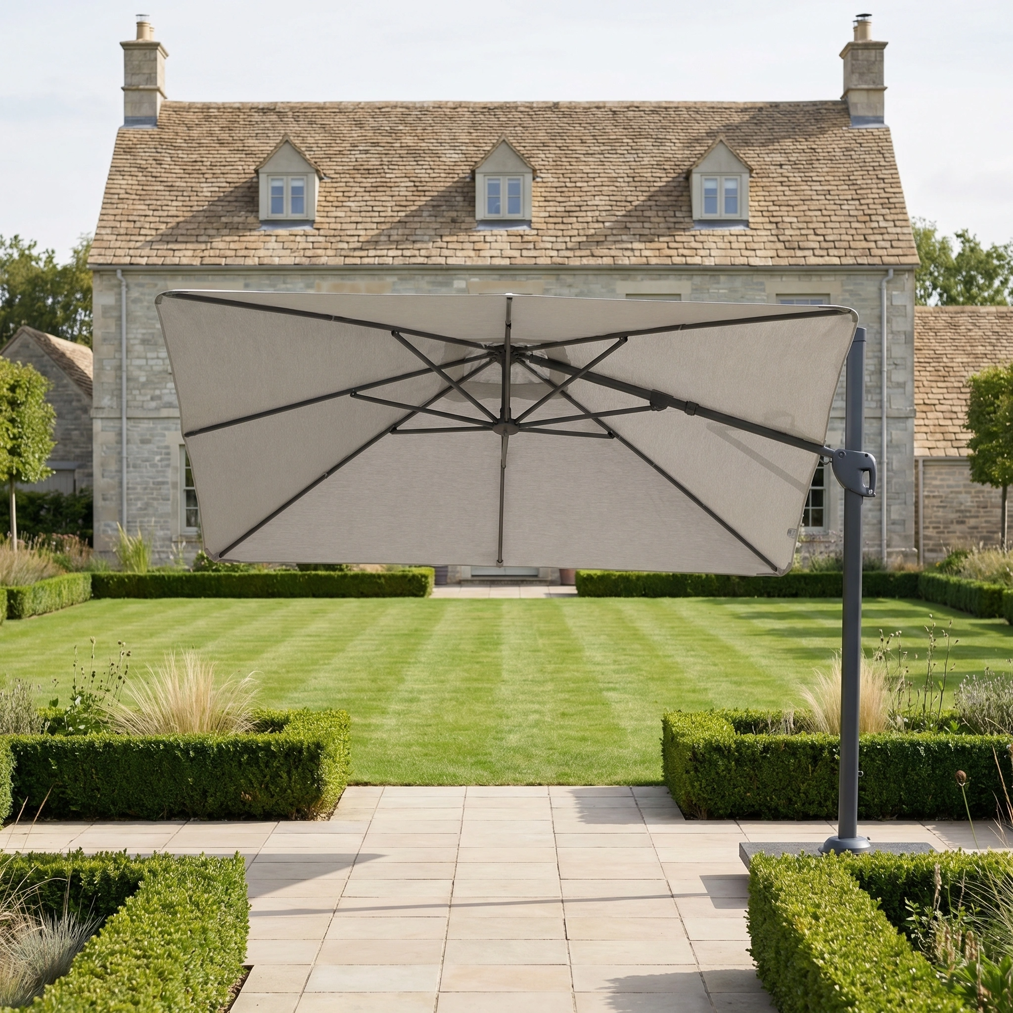 The Nexus T2 Premium 3m Square Cantilever Parasol in Manhattan Grey with 90kg Granite Base, featuring 360-degree rotation, stands on a stone walkway in a manicured garden with a stone house in the background.