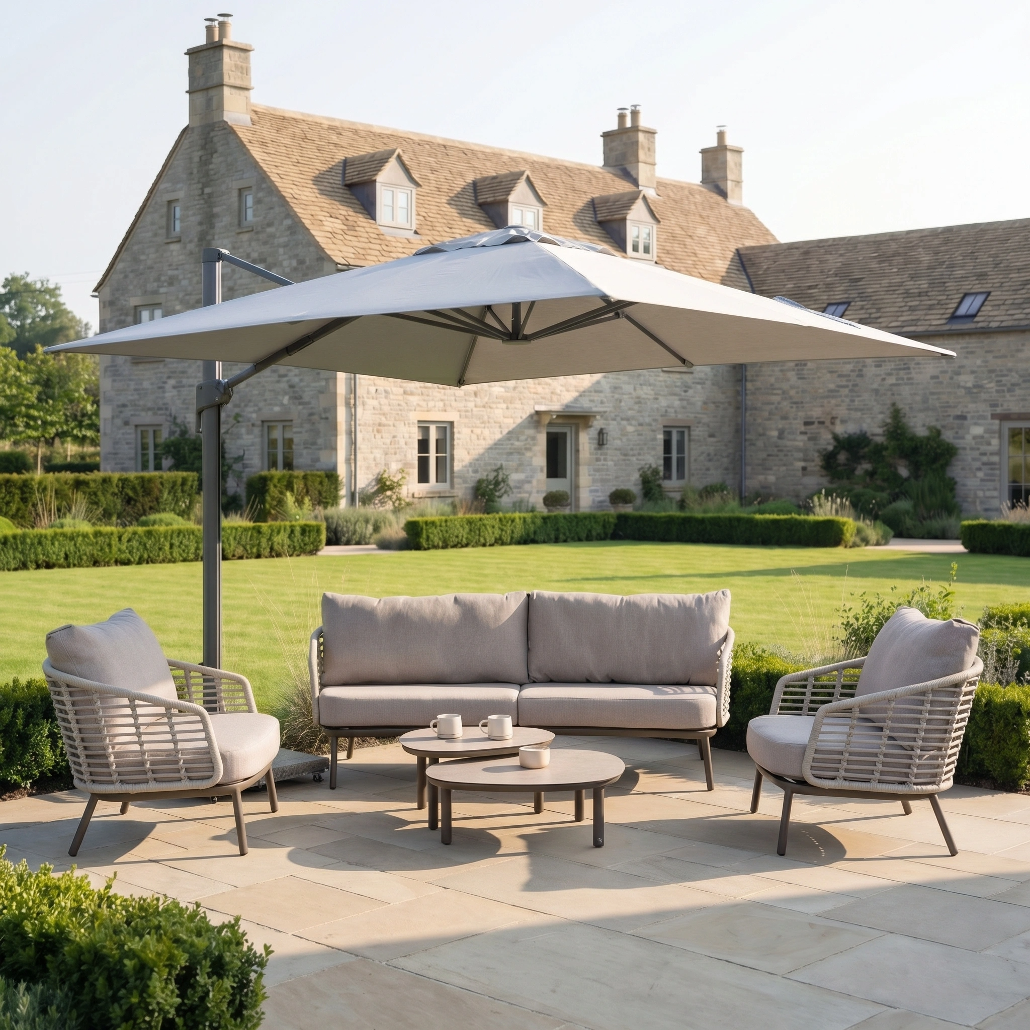 The Nexus T2 Premium 3m Square Cantilever Parasol in Manhattan Grey with a 90kg granite base provides shade over modern patio furniture on a stone terrace, with a stone house and garden visible in the background.