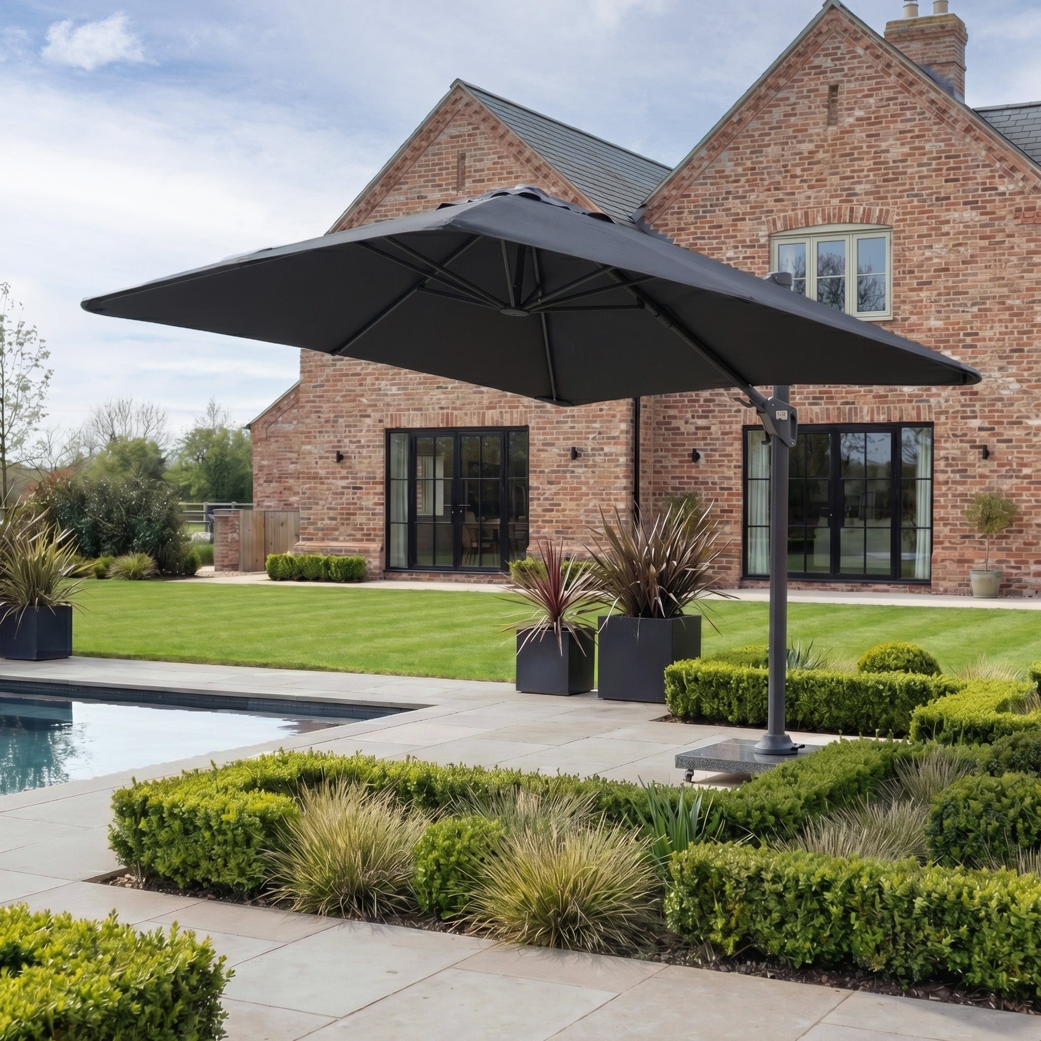 A Nexus T2 Premium 3m Square Cantilever Parasol in Black with a 90kg granite base stands by a pool in a landscaped garden near a brick house.