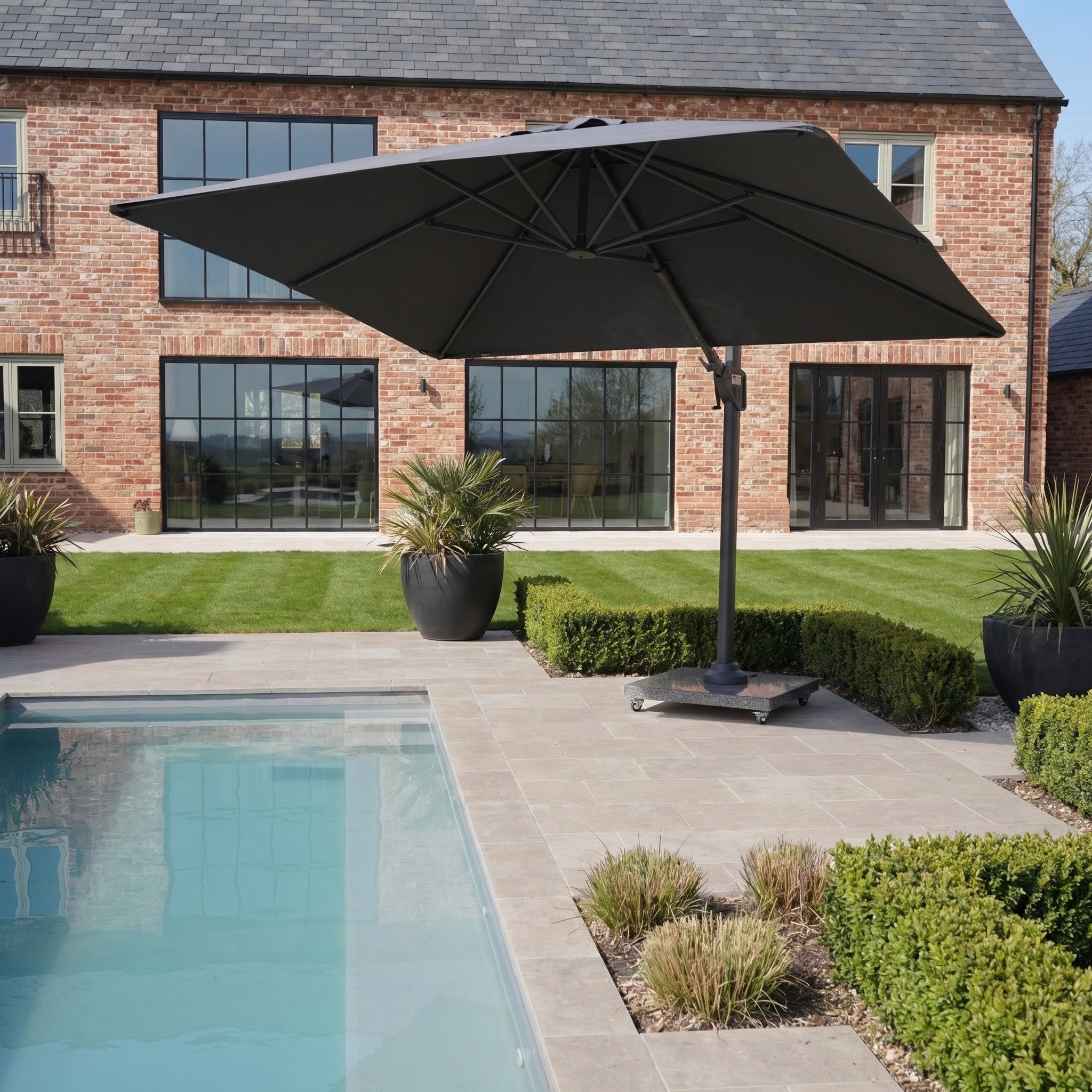 The Nexus T2 Premium 3m Square Cantilever Parasol in Black with 90kg Granite Base offers shade by a modern pool, surrounded by trimmed hedges and a brick house, and features sleek design for contemporary outdoor spaces.
