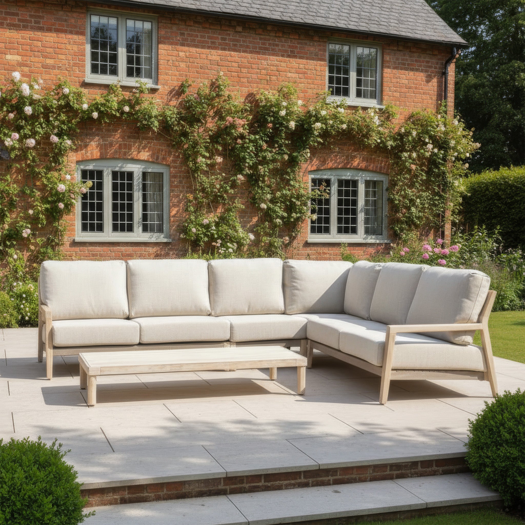 The Noah Corner Sofa Set in Volga Grey and table sit on a patio before a brick house with climbing roses, creating an inviting garden sofa set.