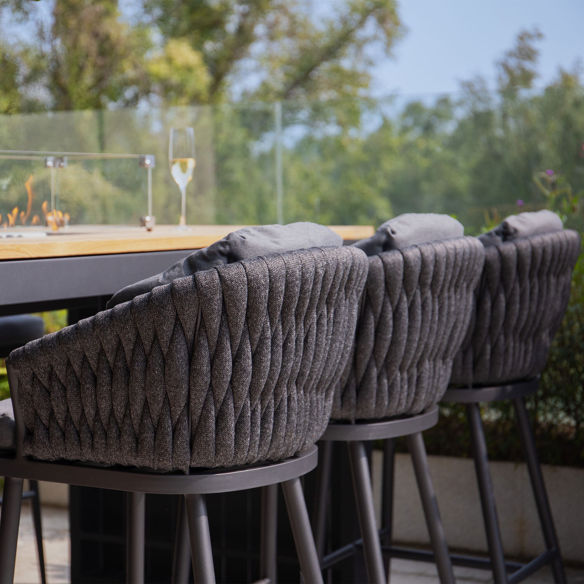 Three cushioned Palma Rope Bar Stools in Grey at an outdoor table in a UV-resistant outdoor living area, with a champagne glass in the background.