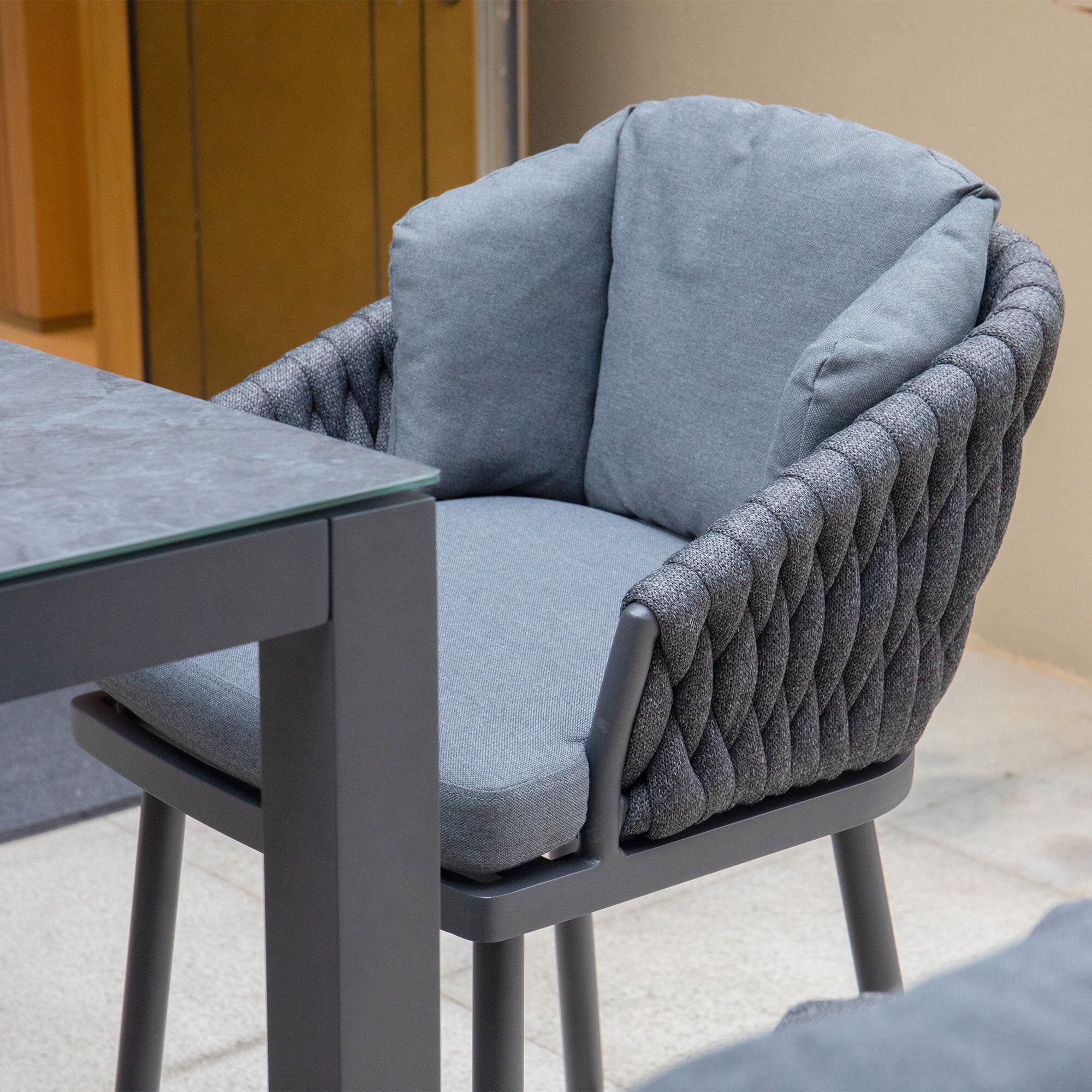The Palma Rope Bar Stool in Grey, featuring a cushioned seat and a woven backrest, pairs perfectly with a table that has a gray marble-like surface, ideal for your outdoor living area.