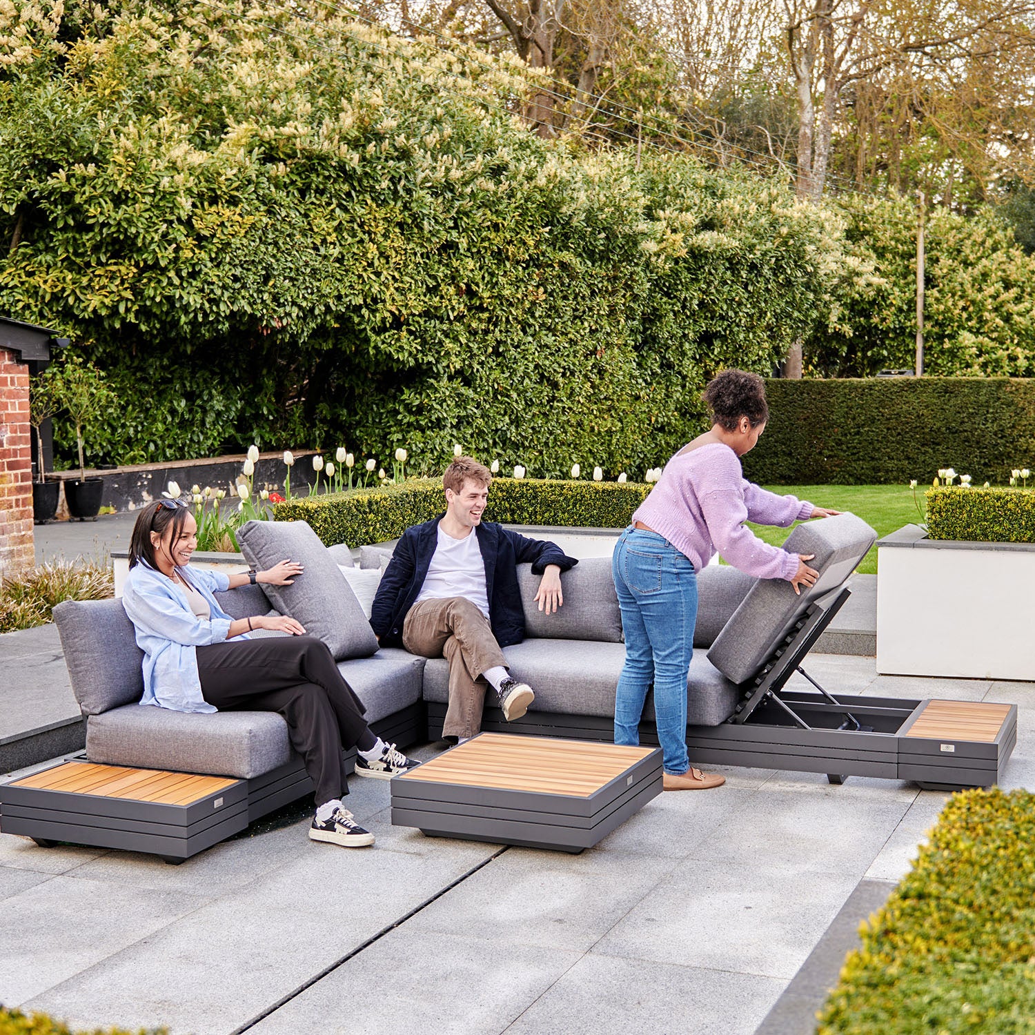 Three people enjoy a Panama Luxury Outdoor L-Shape Corner Group Set in Washed Grey on the patio; one woman lifts a section to reveal storage while others sit and relax.
