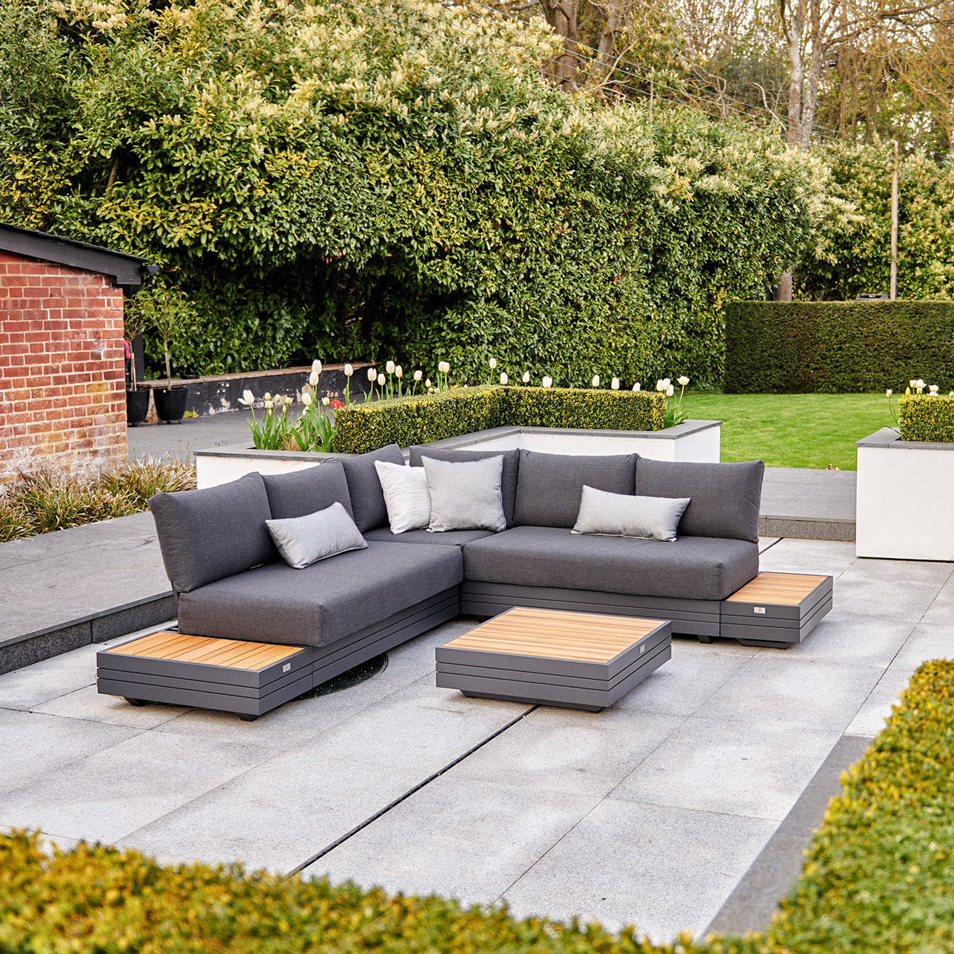 Contemporary outdoor seating area featuring the Panama Luxury Outdoor Corner Group Set in Charcoal, wooden tables crafted from sustainable teak, and green hedges in the background.