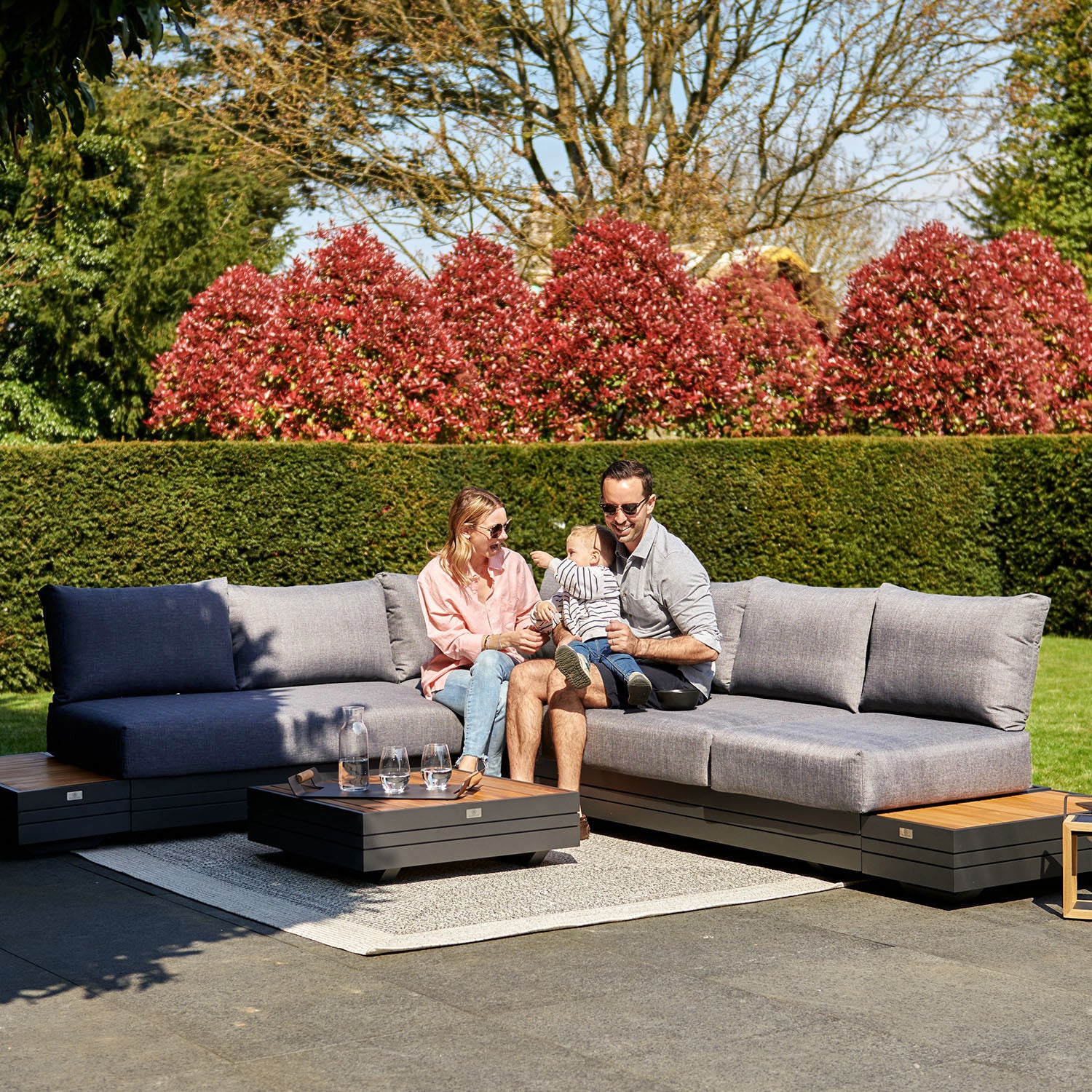 A couple sits on an outdoor sofa with their baby, surrounded by greenery and red-leafed bushes in the background. Their elegant Panama Luxury Outdoor L-Shape Corner Group Set in Washed Grey exemplifies luxury outdoor furniture and adds a touch of sophistication to the serene setting.