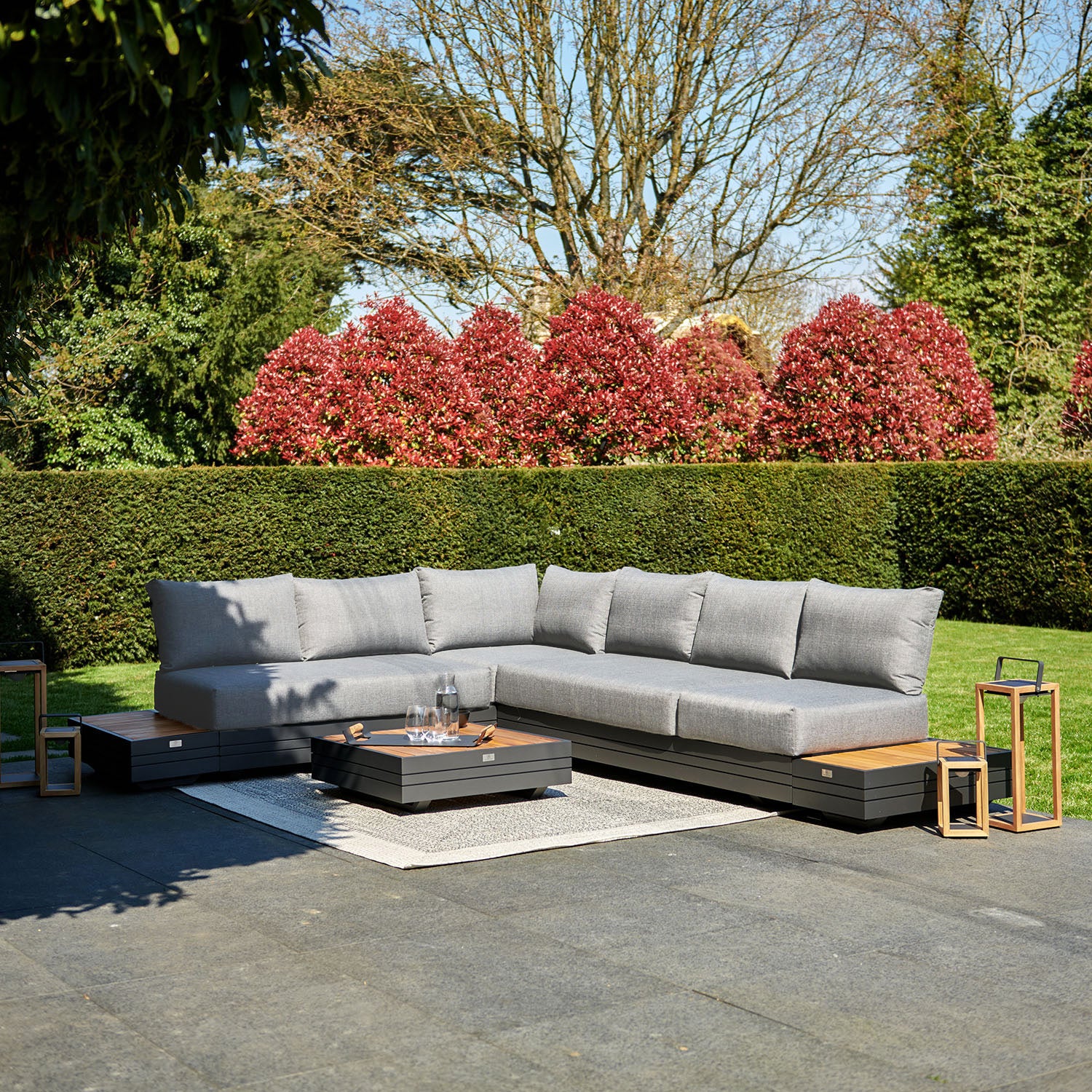 An outdoor patio showcases the Panama Luxury Outdoor L-Shape Corner Group Set in Washed Grey, featuring a stylish sectional sofa, a low coffee table made of sustainable teak, and vibrant red foliage in the background on a sunny day.