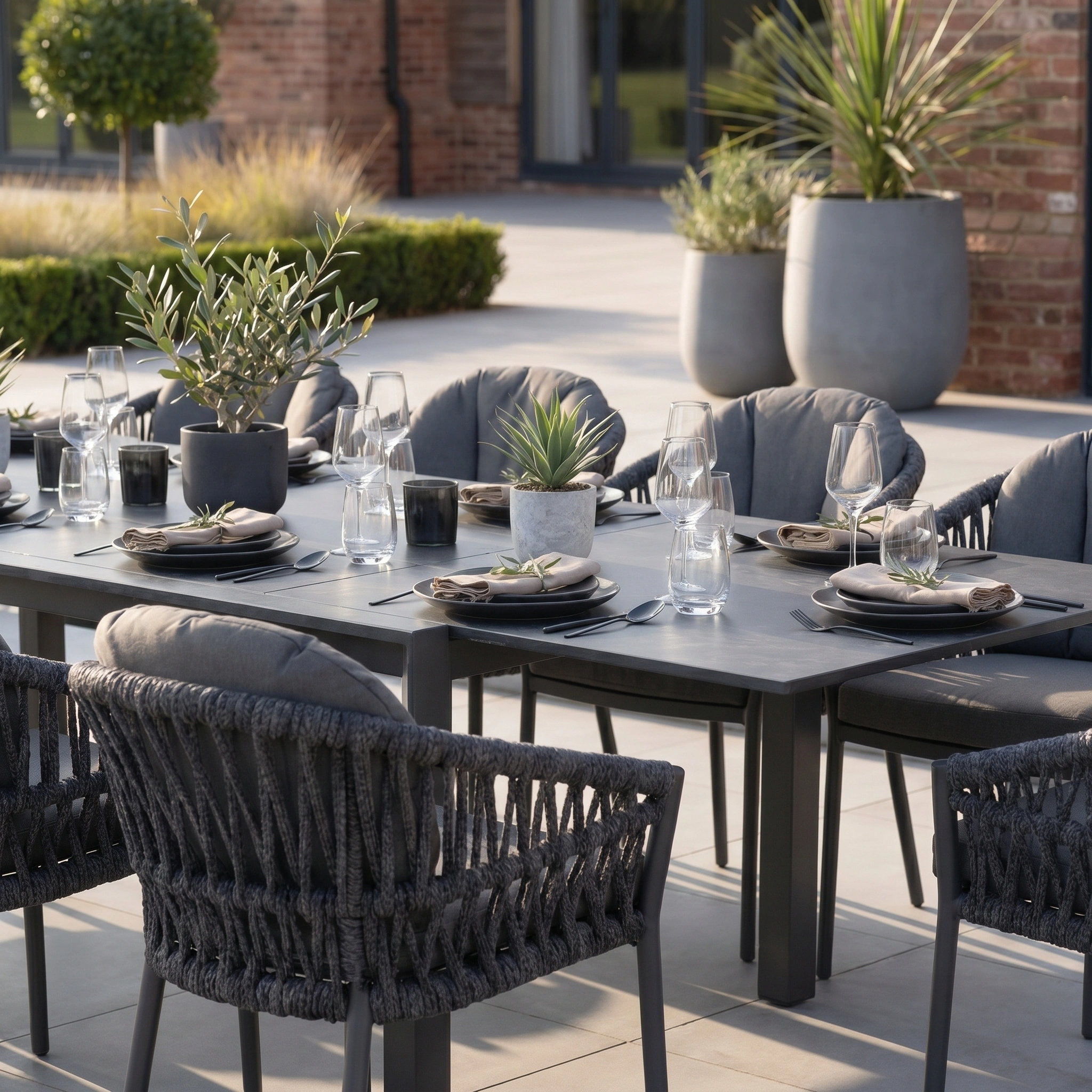 The Pello 10 Seat Extending Dining Set with Ceramic Table in Grey features modern chairs, elegant place settings, glassware, and potted plants—ideal for stylish outdoor gatherings on a spacious patio.