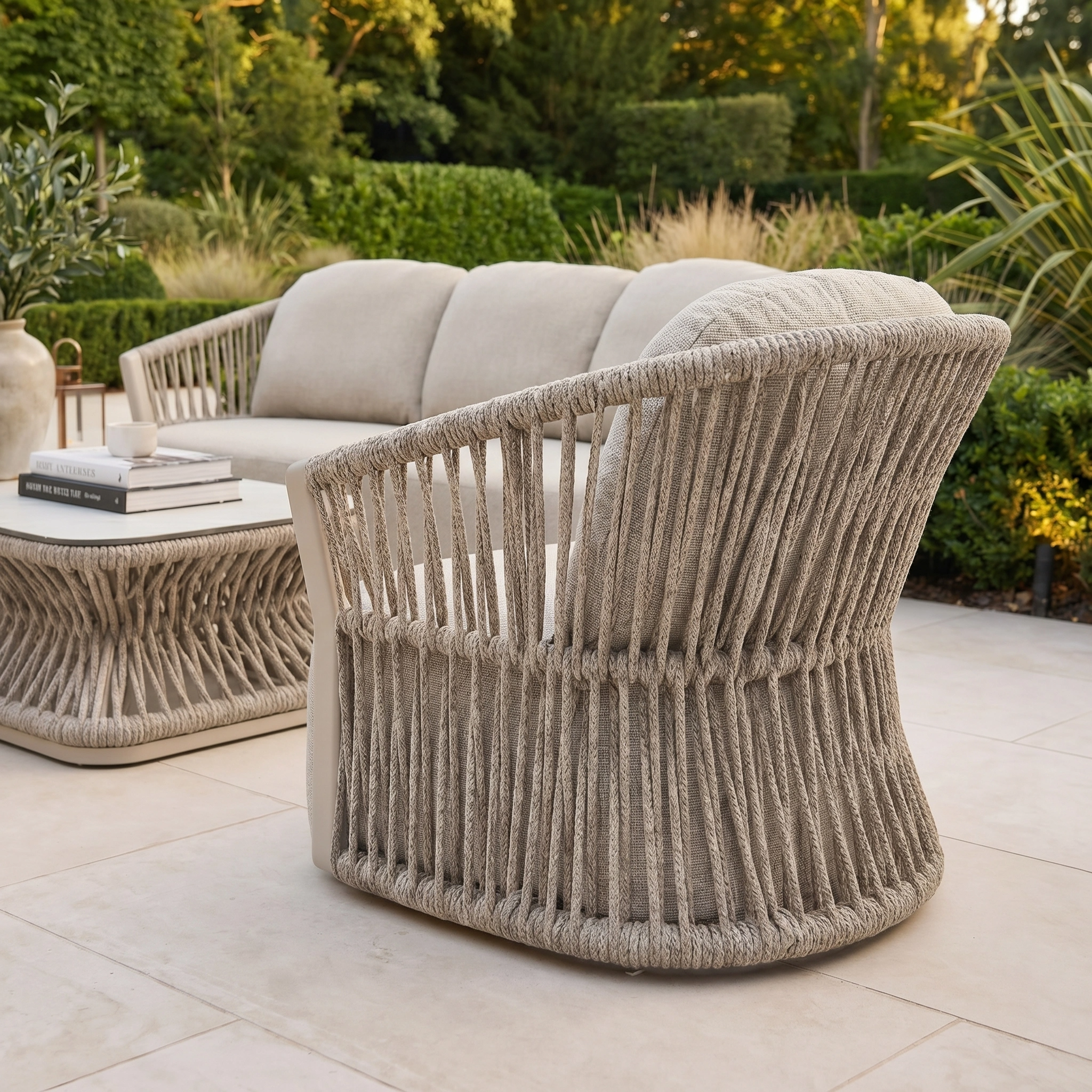 Outdoor patio featuring the Pello 3 Seat Sofa Set in Fawn with woven rope design, quick dry cushions, and a coffee table with books, all framed by lush greenery.