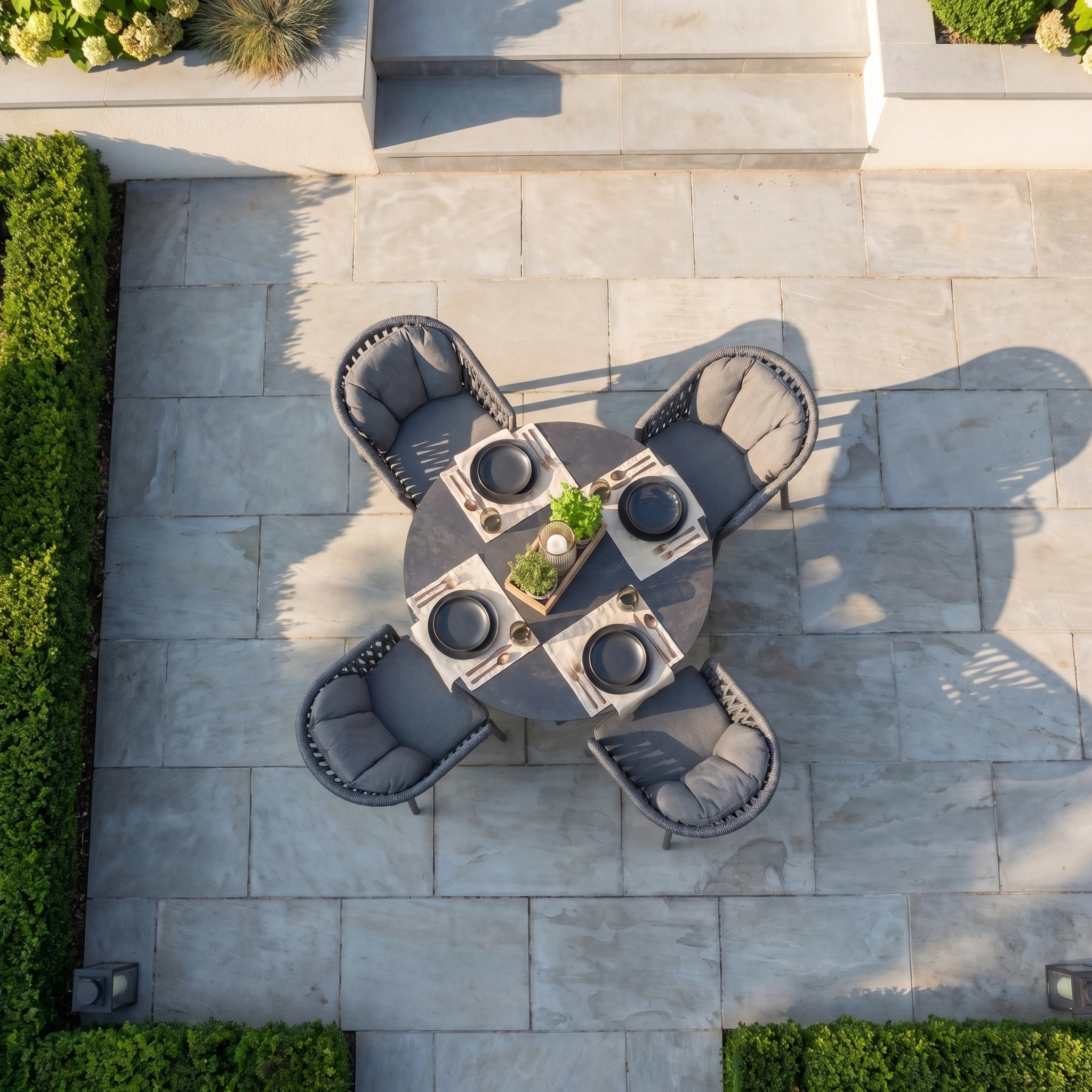 The Pello 4 Seat Round Dining Set with Albatross Table in Grey offers a cozy spot for four, perfect on stone tiles and surrounded by green hedges.