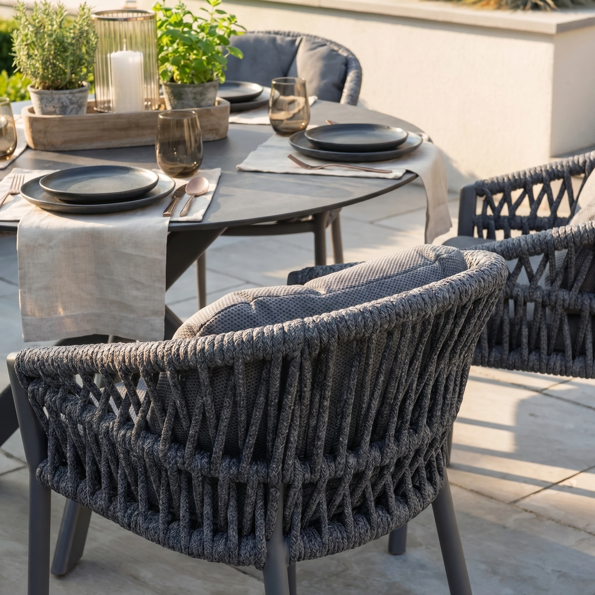 The Pello 4 Seat Round Dining Set with Albatross Table in Grey features woven chairs, black plates, glasses, and potted plants as a centerpiece—perfect for stylish outdoor dining.