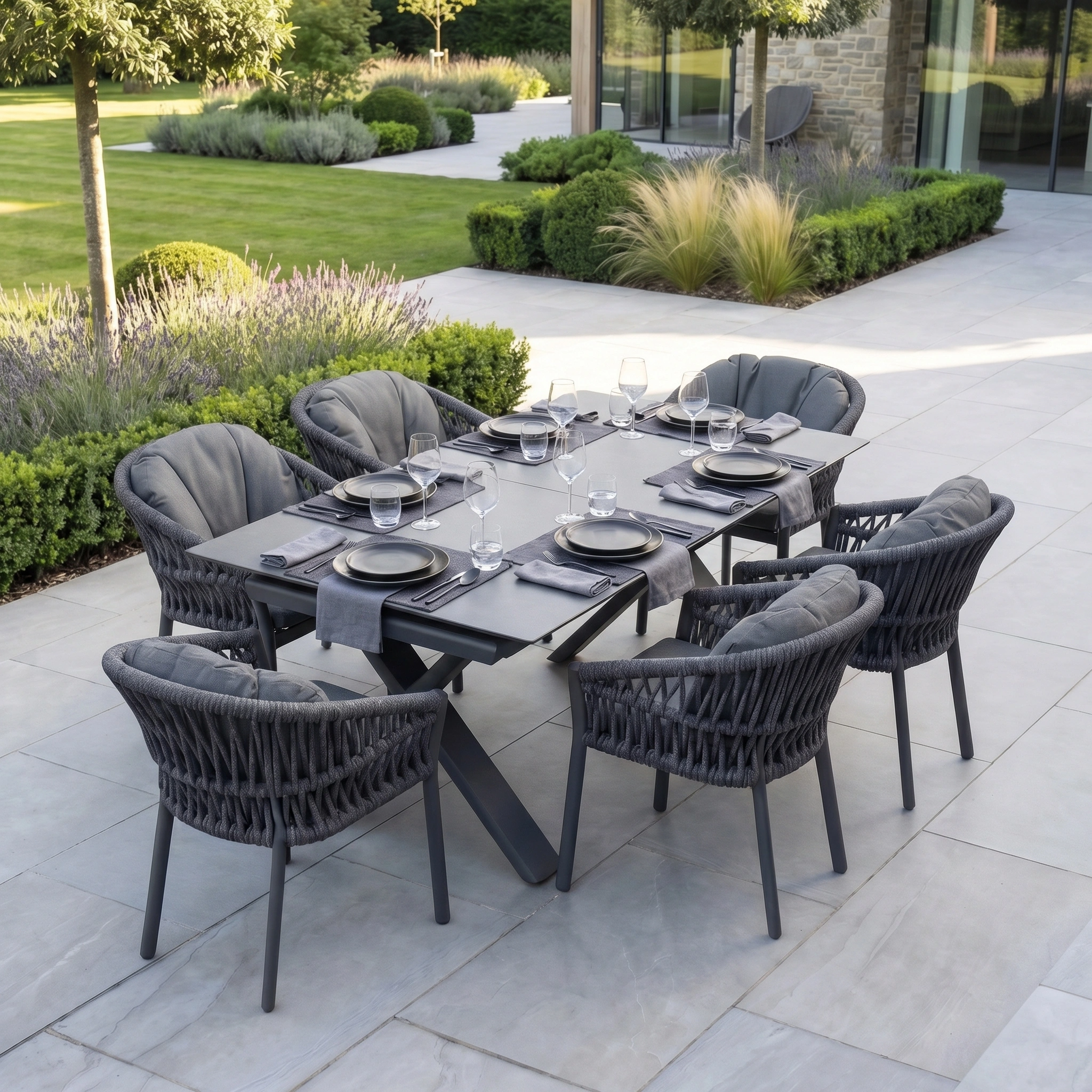 The Pello 6 Seat Dining Set with Albatross Extending Table in Grey provides comfortable outdoor dining with stylish dark chairs and elegant tableware, arranged on a patio amidst lush garden greenery.