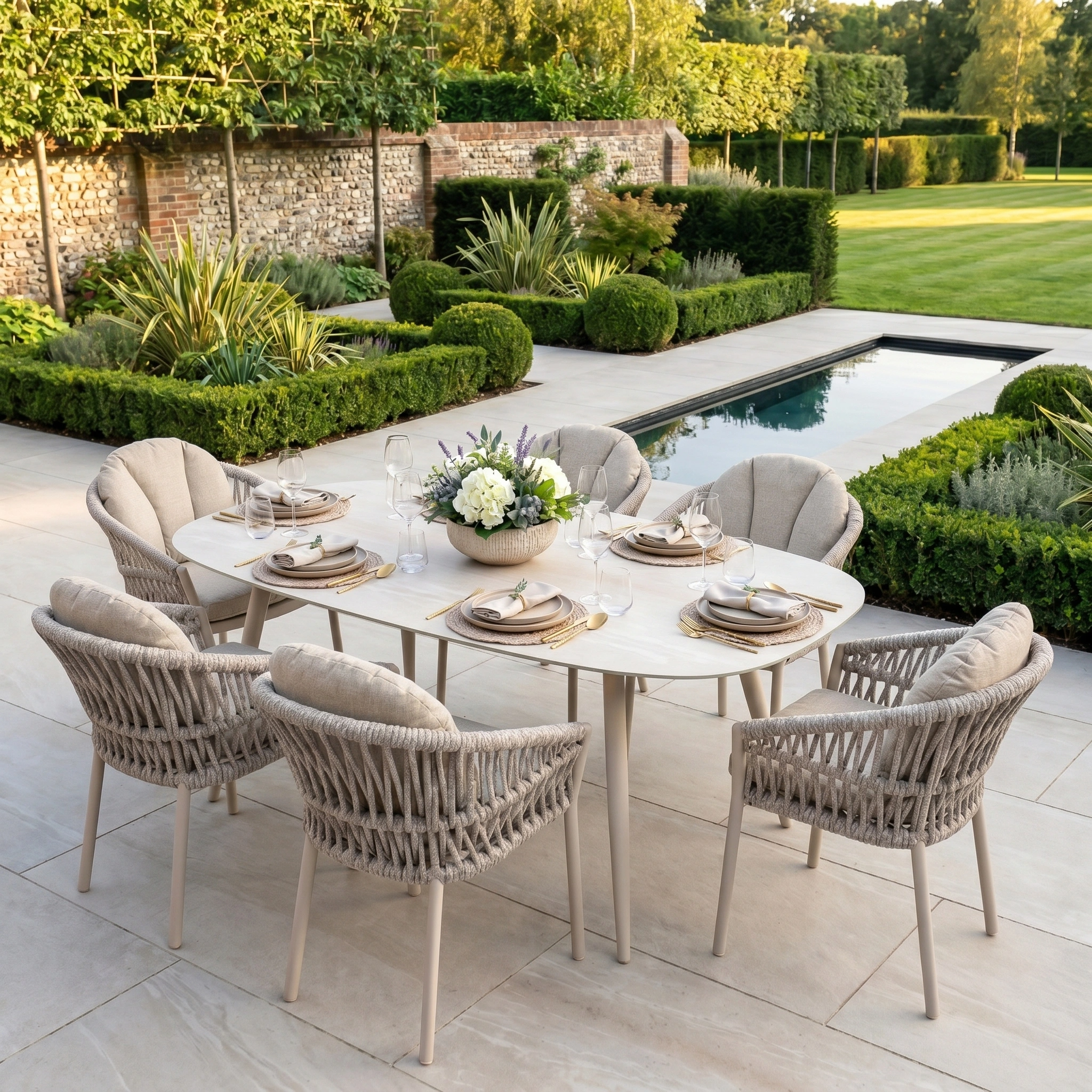 The Pello 6 Seat Oval Dining Set with Ceramic Table in Fawn, arranged poolside amid lush gardens, offers weather-resistant style—perfect for elegant outdoor dining.
