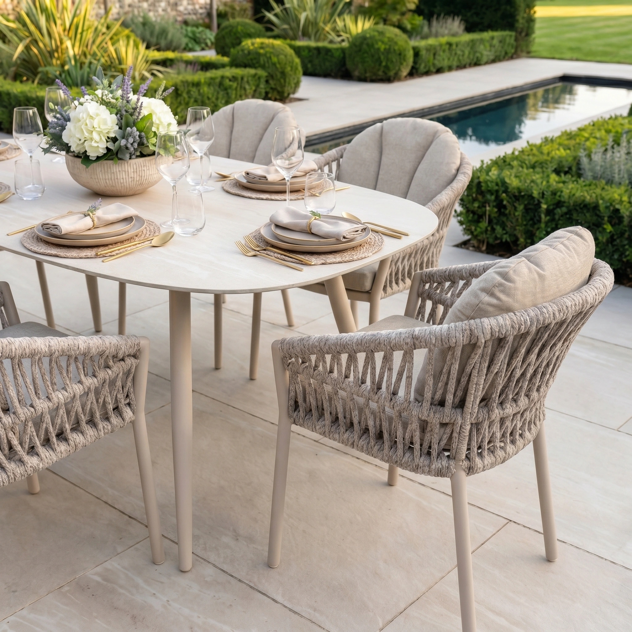 The Pello 6 Seat Oval Dining Set with Ceramic Table in Fawn features elegant tableware and wicker chairs, creating a stylish, weather-resistant outdoor dining area beside your landscaped pool and garden.