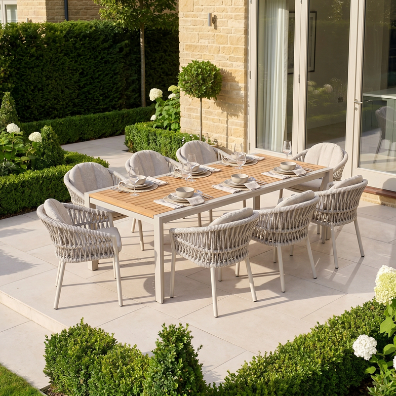 The Pello 8 Seat Extending Dining Set with Teak Table in Fawn is a weather-resistant outdoor ensemble, ideal for gatherings, featuring a teak table and eight chairs set on a patio surrounded by greenery and flowers.
