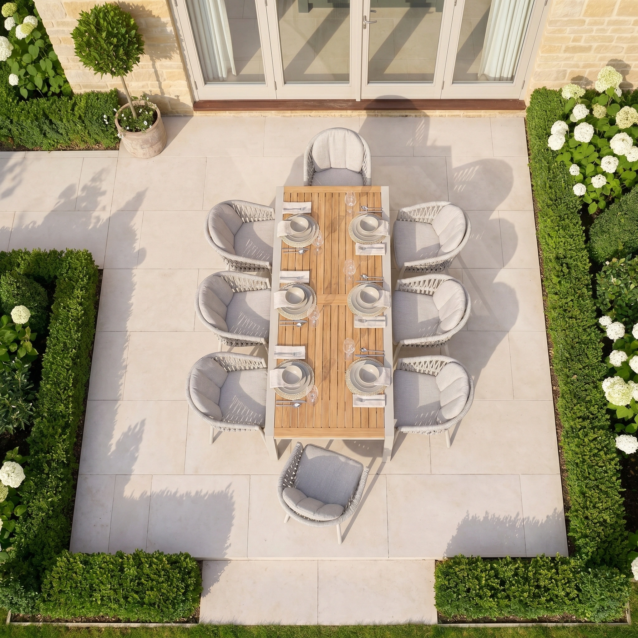The Pello 8 Seat Extending Dining Set with Teak Table in Fawn is a weather-resistant outdoor furniture choice, perfect for gatherings on a tiled patio surrounded by green hedges and white flowers.