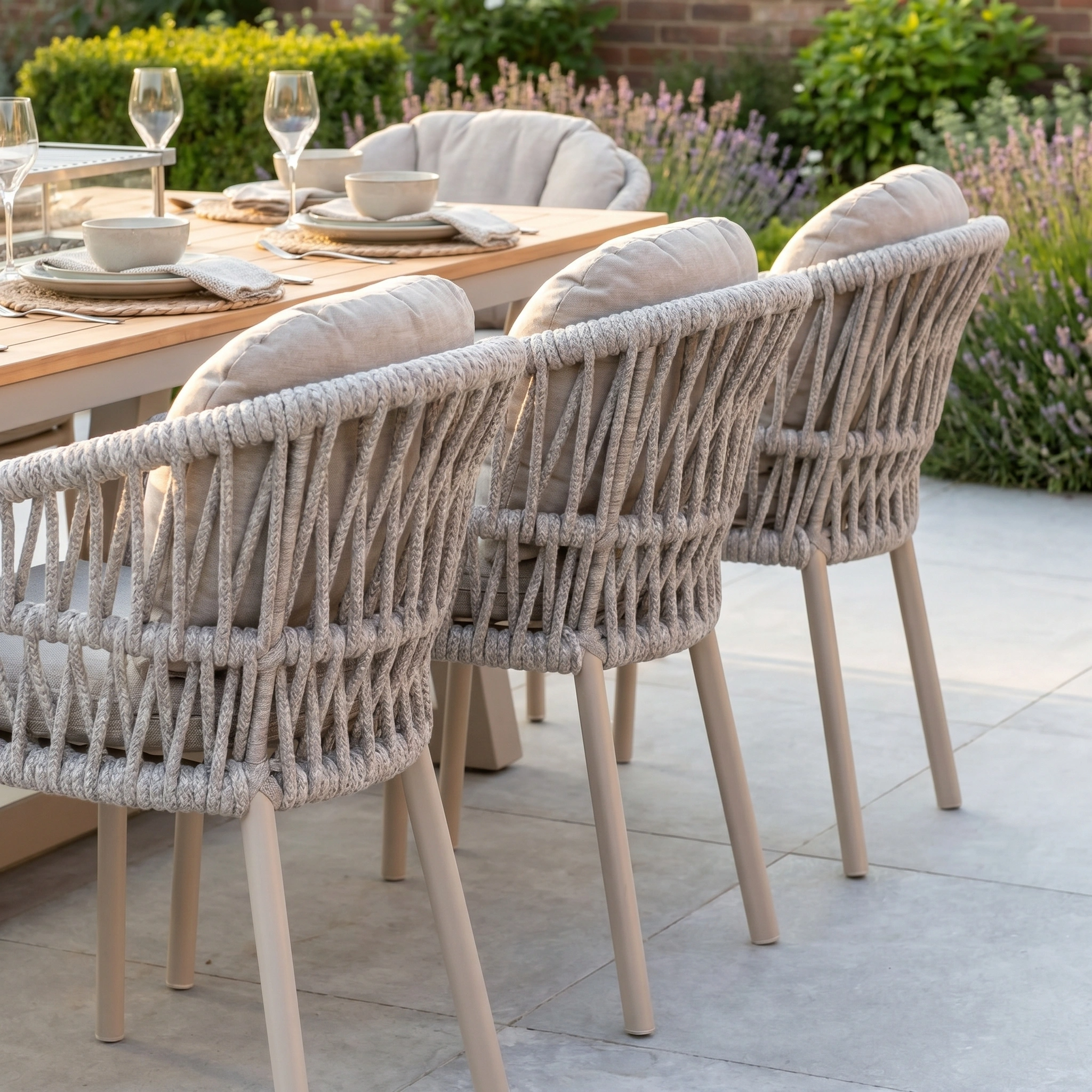The Pello 8 Seat Rectangular Teak Firepit Dining Set in Fawn features cushioned rope chairs and a teak table, elegantly set with plates and glassware, perfect for outdoor dining in a lush garden setting.