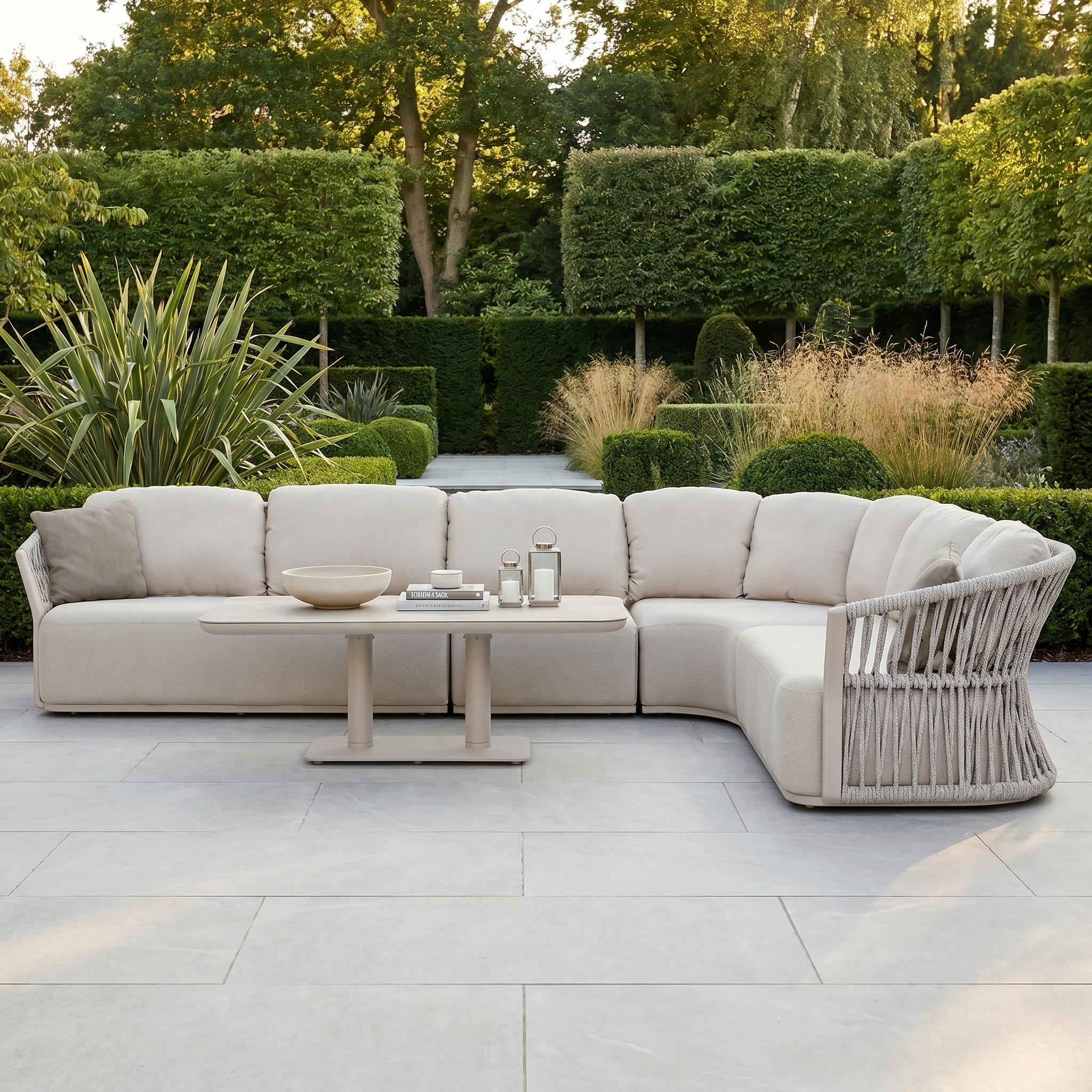 The Pello Deluxe Curved Sofa Set with Altura Rising Table in Fawn features quick-dry foam cushions, ideal for outdoor living in a landscaped garden with lush greenery and stone tiles.