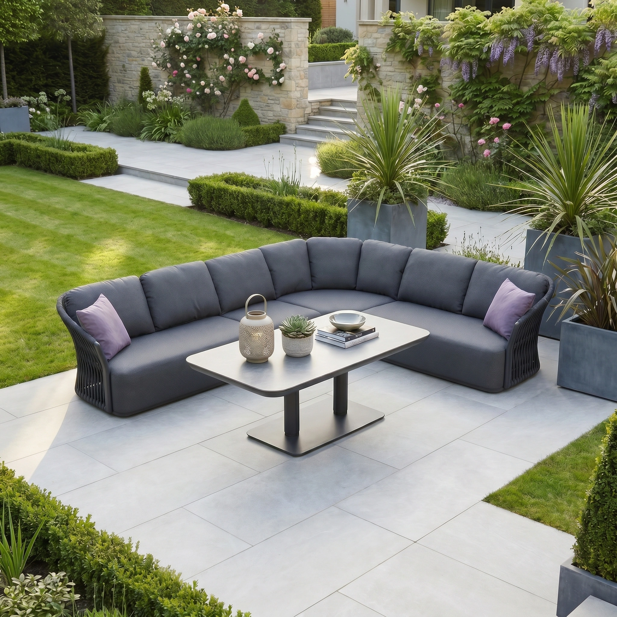 Pello Deluxe Curved Sofa Set with Altura Rising Table in Grey—stylish outdoor furniture with cushions and table, ideal for relaxing or entertaining on your patio amid a lush green garden and flowers.
