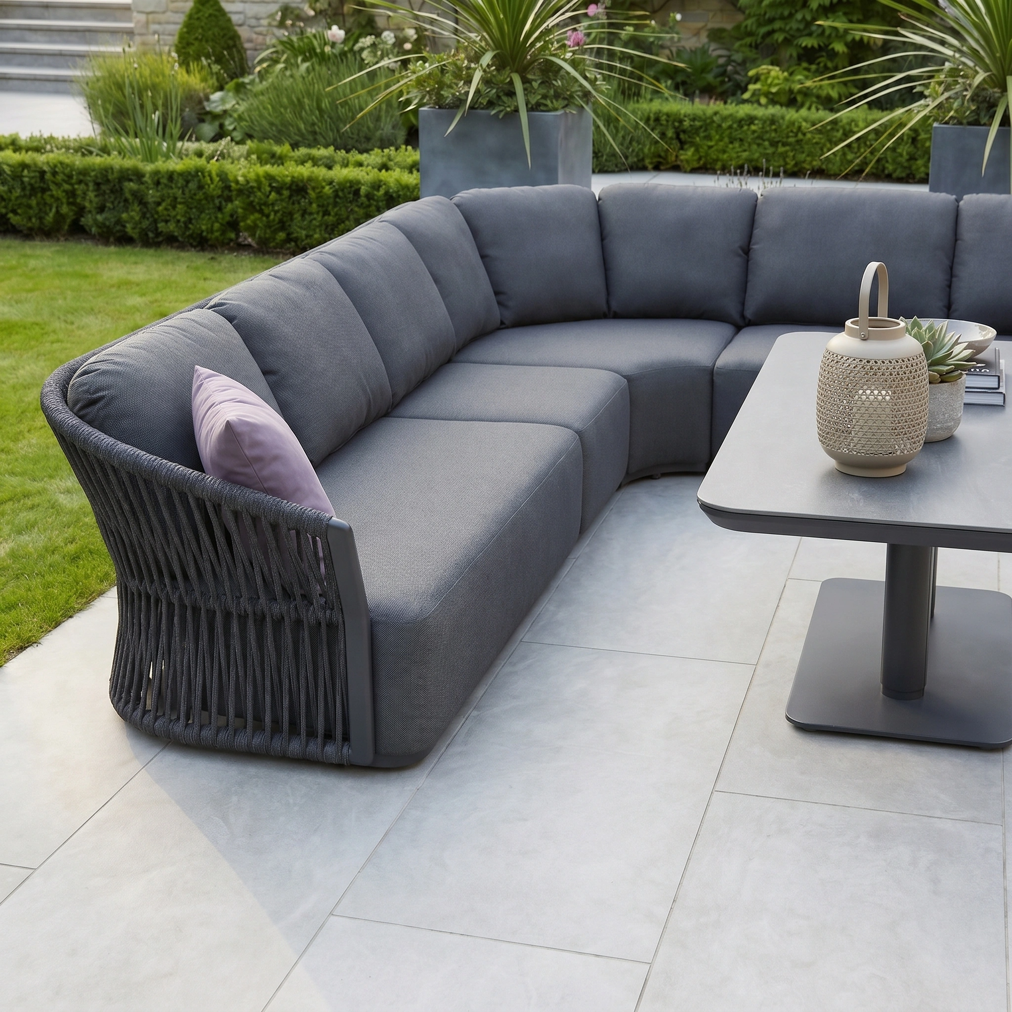 The Pello Deluxe Curved Sofa Set with Altura Rising Table in Grey sits on a tiled patio by the grass, with lush plants in the background for a relaxing garden vibe.