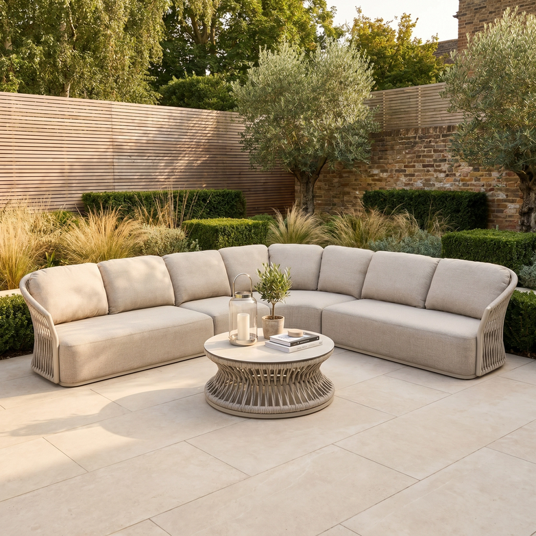 The Pello Deluxe Curved Sofa Set with Round Coffee Table in Fawn features weather-resistant cushions—ideal for stylish outdoor relaxation on your patio, surrounded by greenery and wooden fencing.