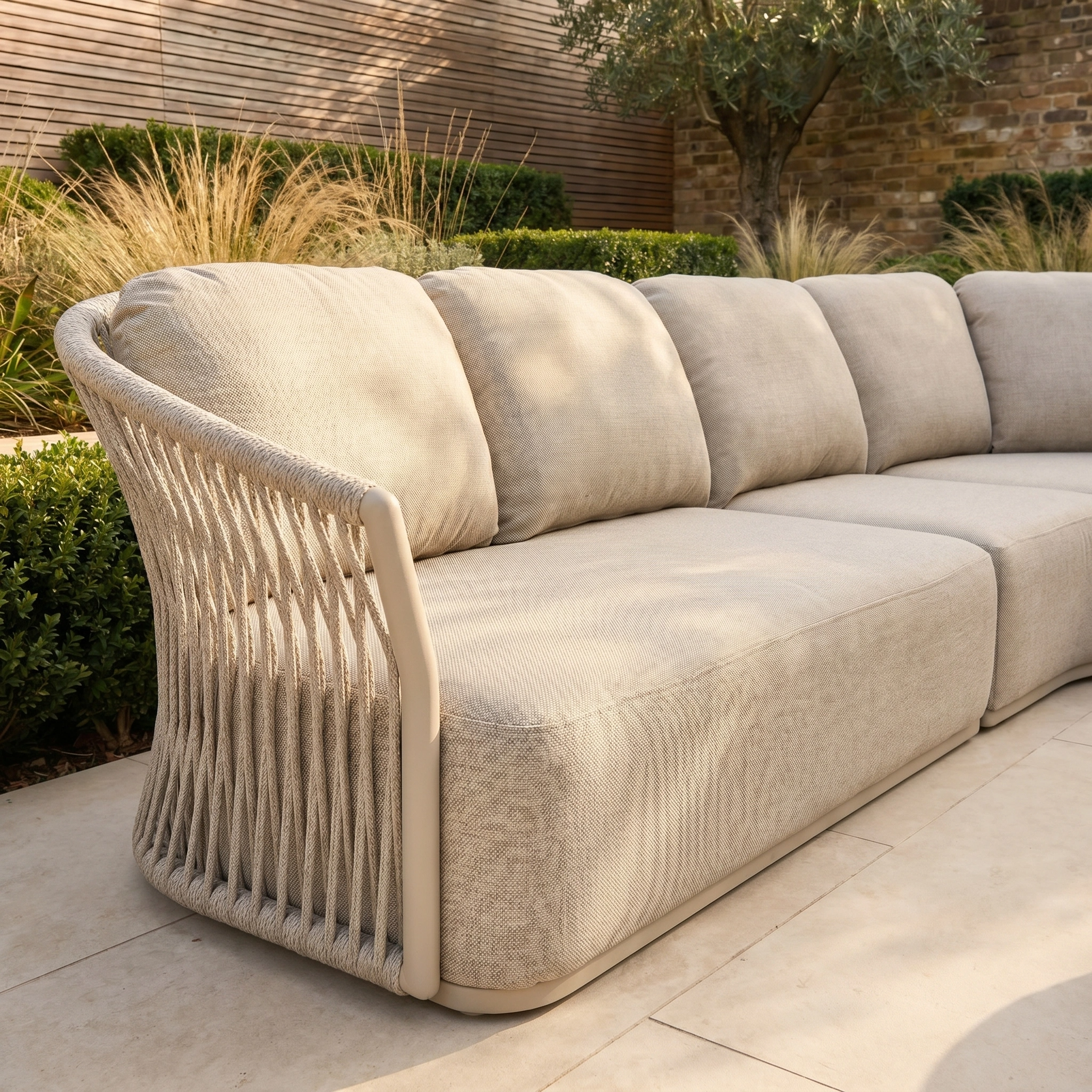 The Pello Deluxe Curved Sofa Set with Round Coffee Table in Fawn features rope-detailed armrests and weather-resistant cushions, ideal for outdoor use on a stone patio surrounded by plants.