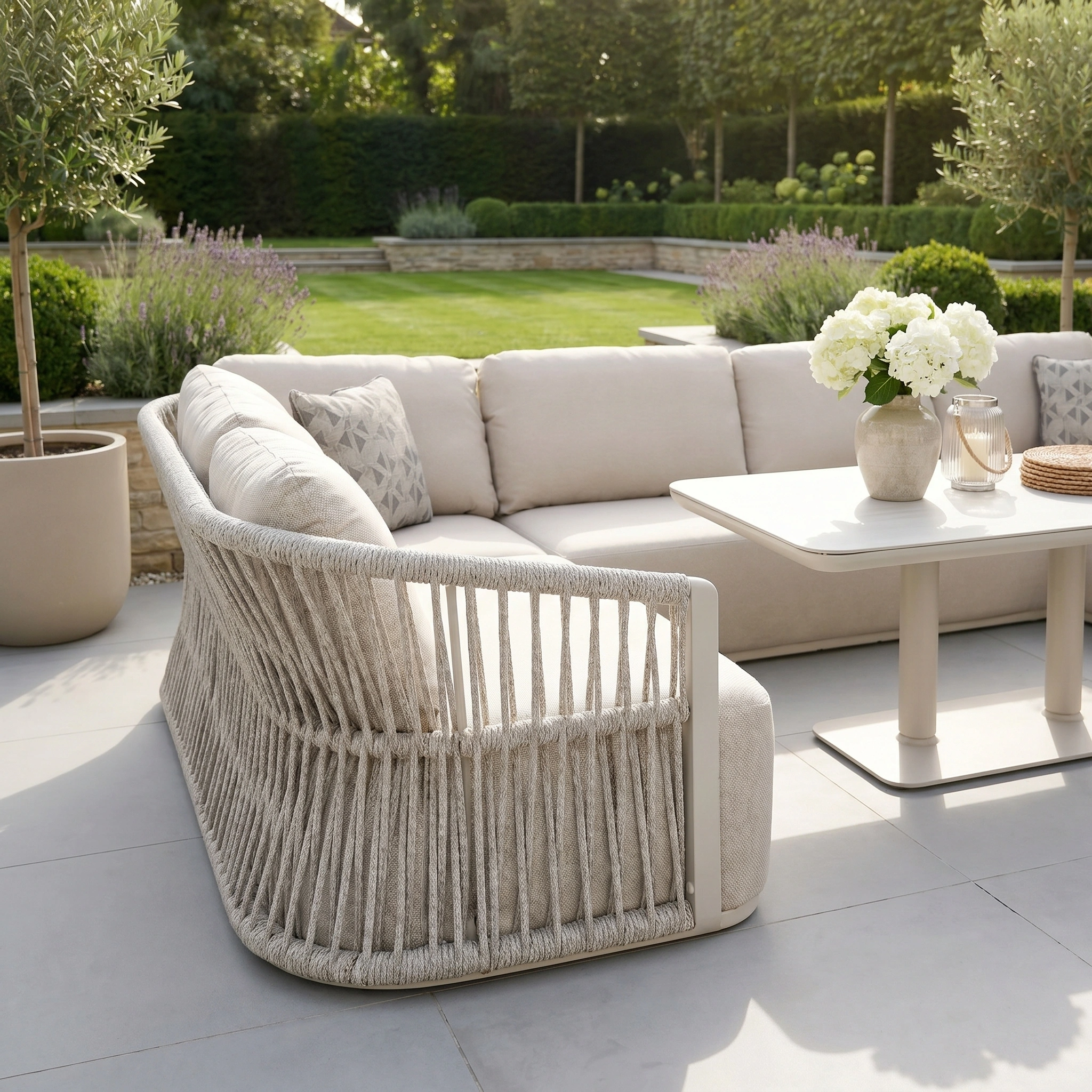 Create an inviting patio with the Pello U-Shape Corner Sofa Set in Fawn, paired with an Altura Rising Table and white cushions, perfect for relaxing or entertaining. Add a vase of white flowers to complete this stylish garden setup.