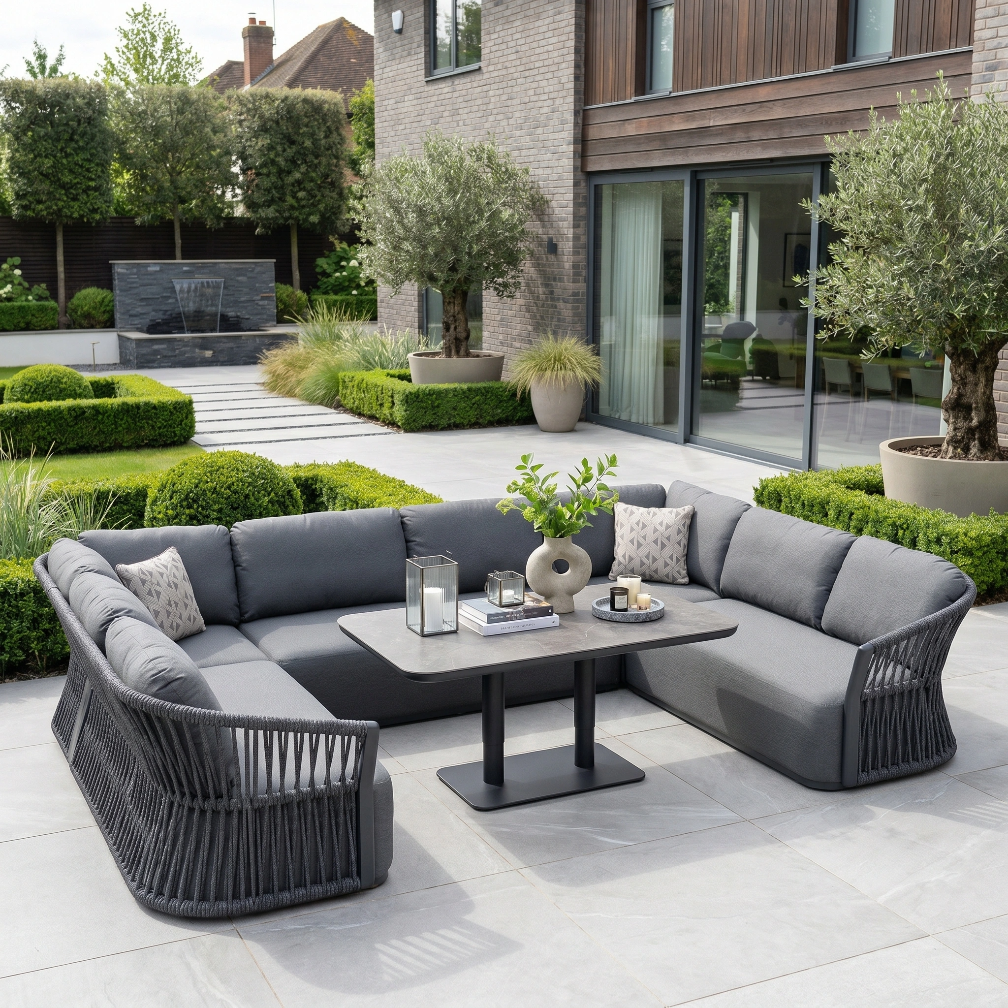 Modern outdoor patio featuring the Pello U-Shape Corner Sofa Set with Altura Rising Table in Grey, plush cushions, and lush plants in a beautifully landscaped garden.
