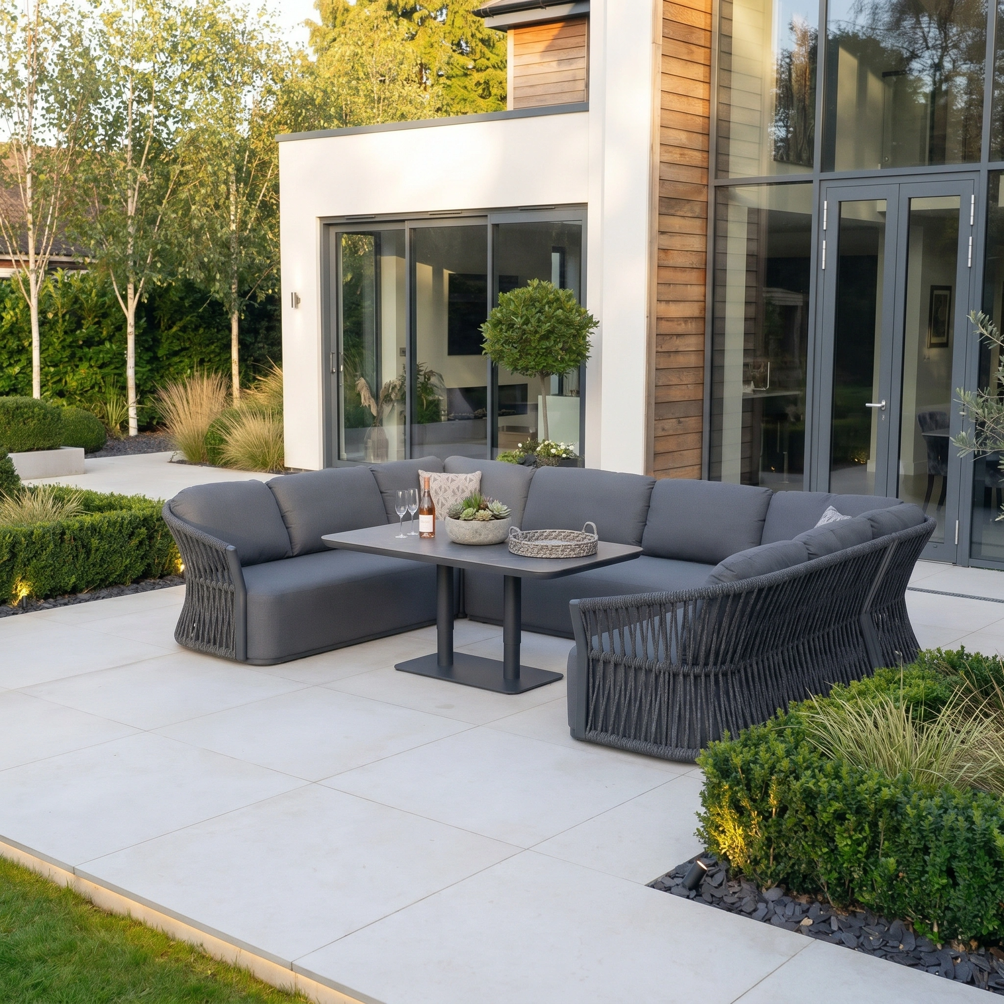 Modern outdoor patio featuring the Pello U-Shape Corner Sofa Set with Altura Rising Table in Grey and lush greenery beside a contemporary house.