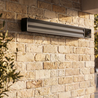 The PergoSTET 2400W Wifi & Bluetooth Heater in Black by Harbour Lifestyle is wall-mounted on an exterior brick wall near greenery—ideal for cosy outdoor evenings under pergolas or patios.