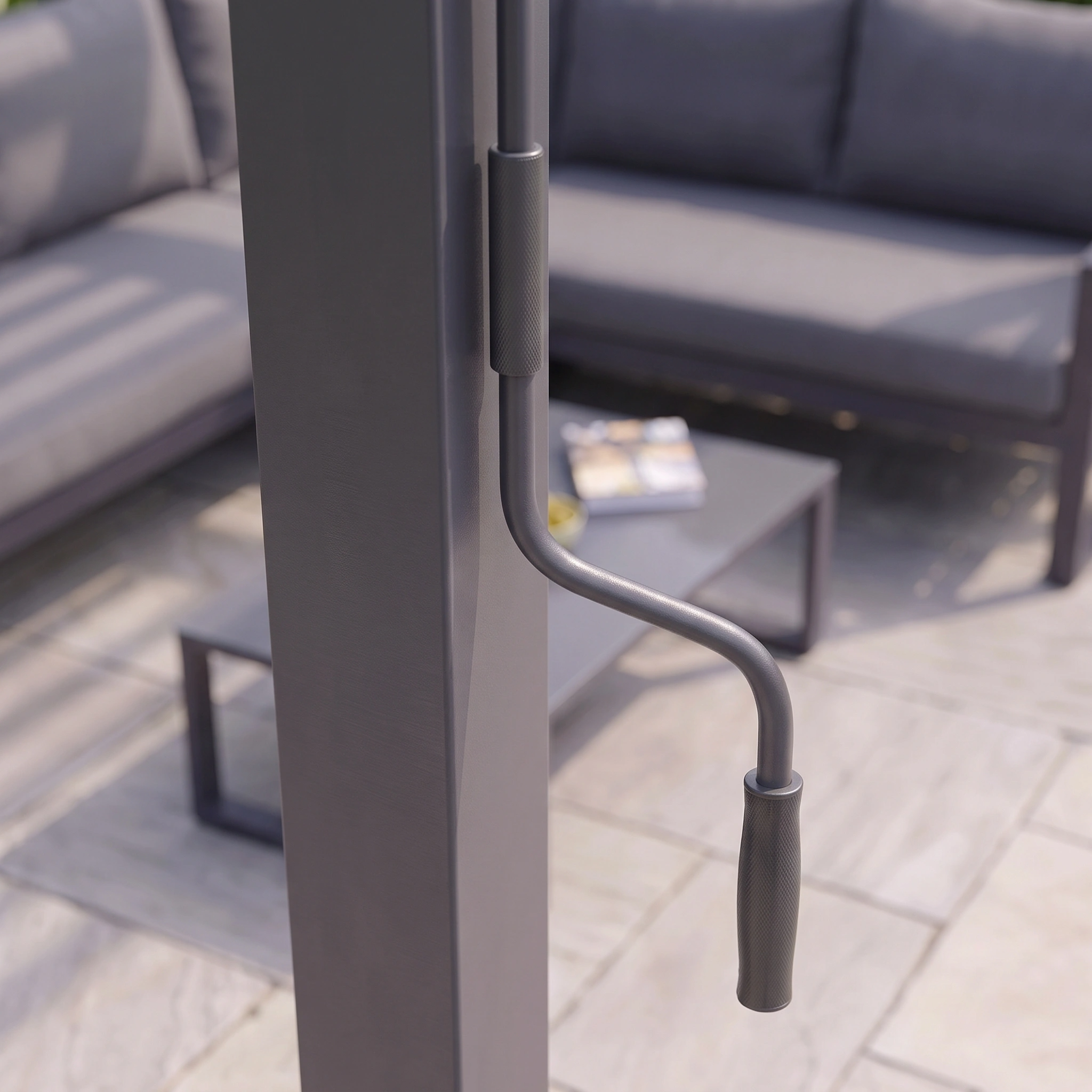 Close-up of a crank handle on a patio umbrella, with an outdoor sofa and table in the background, alongside the PergoSTET Lite 2.4m x 3m Metal Rectangular Pergola in Grey, which features modern tilting louvres for customizable shade.