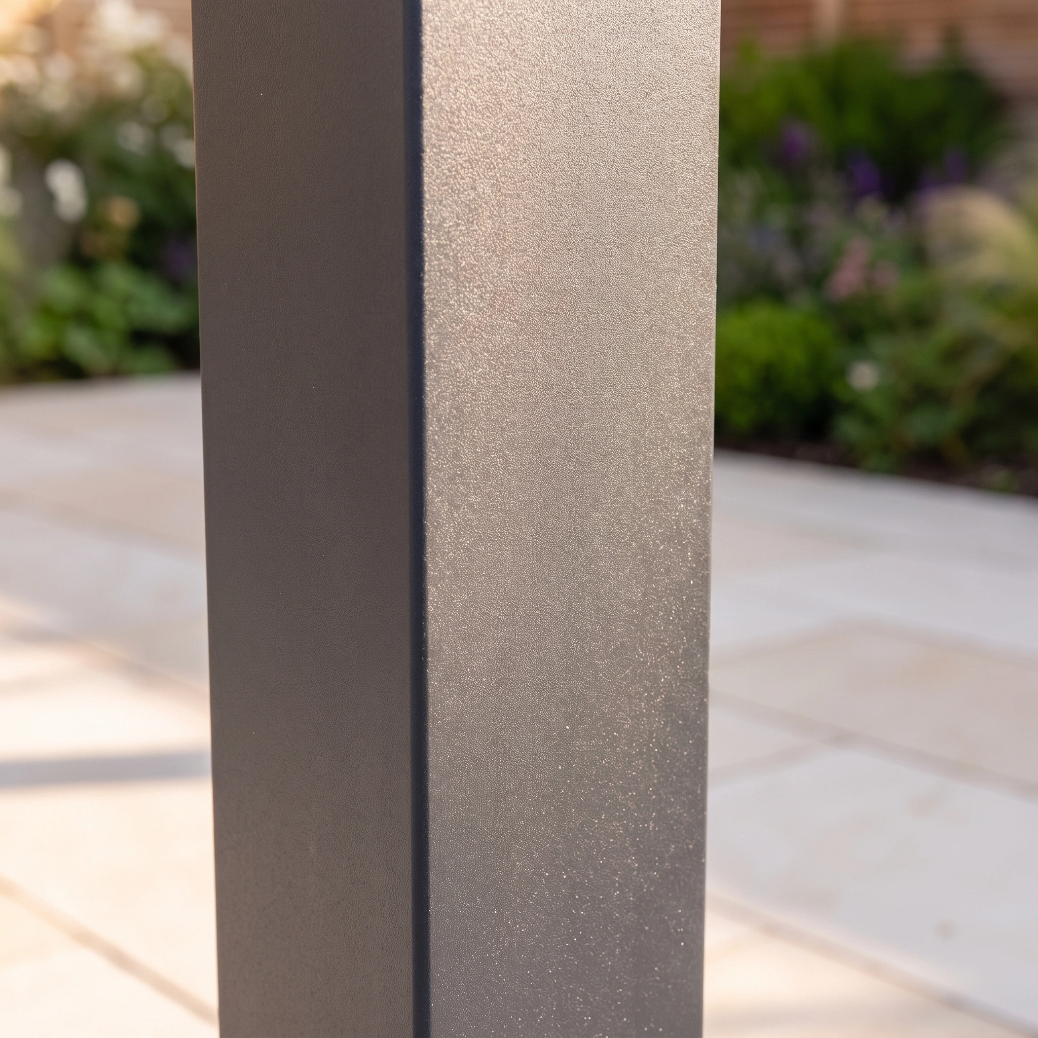 Close-up of a matte gray metal post outdoors, part of the PergoSTET Lite 2.4m x 3m Metal Rectangular Pergola in Grey, with blurred paving tiles and green plants in the background.