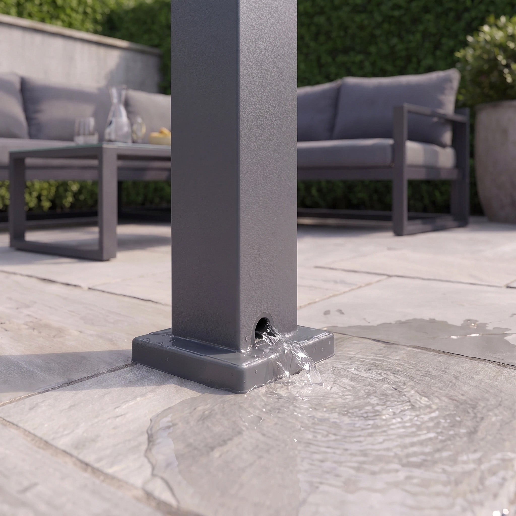 Water flows from the base of a PergoSTET Lite 2.4m x 3m Metal Rectangular Pergola in Grey on a patio with furniture in the background, demonstrating the efficiency of its integrated drainage system.