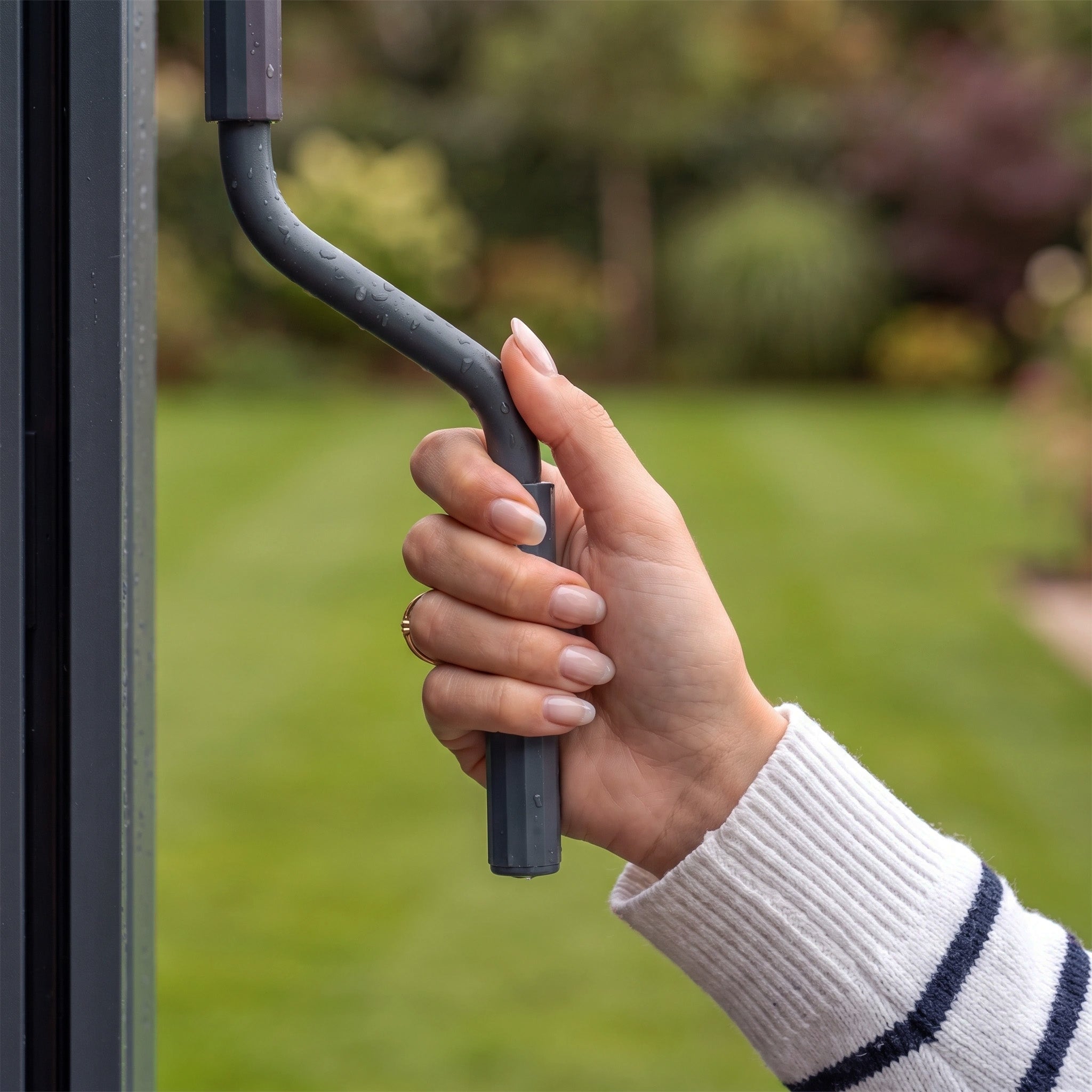 A hand with manicured nails turns the handle on the sleek PergoSTET Lite 2.4m x 3m Metal Rectangular Pergola in Grey, adjusting its tilting louvres for customizable outdoor shade, while a garden and lawn are visible in the blurred background.